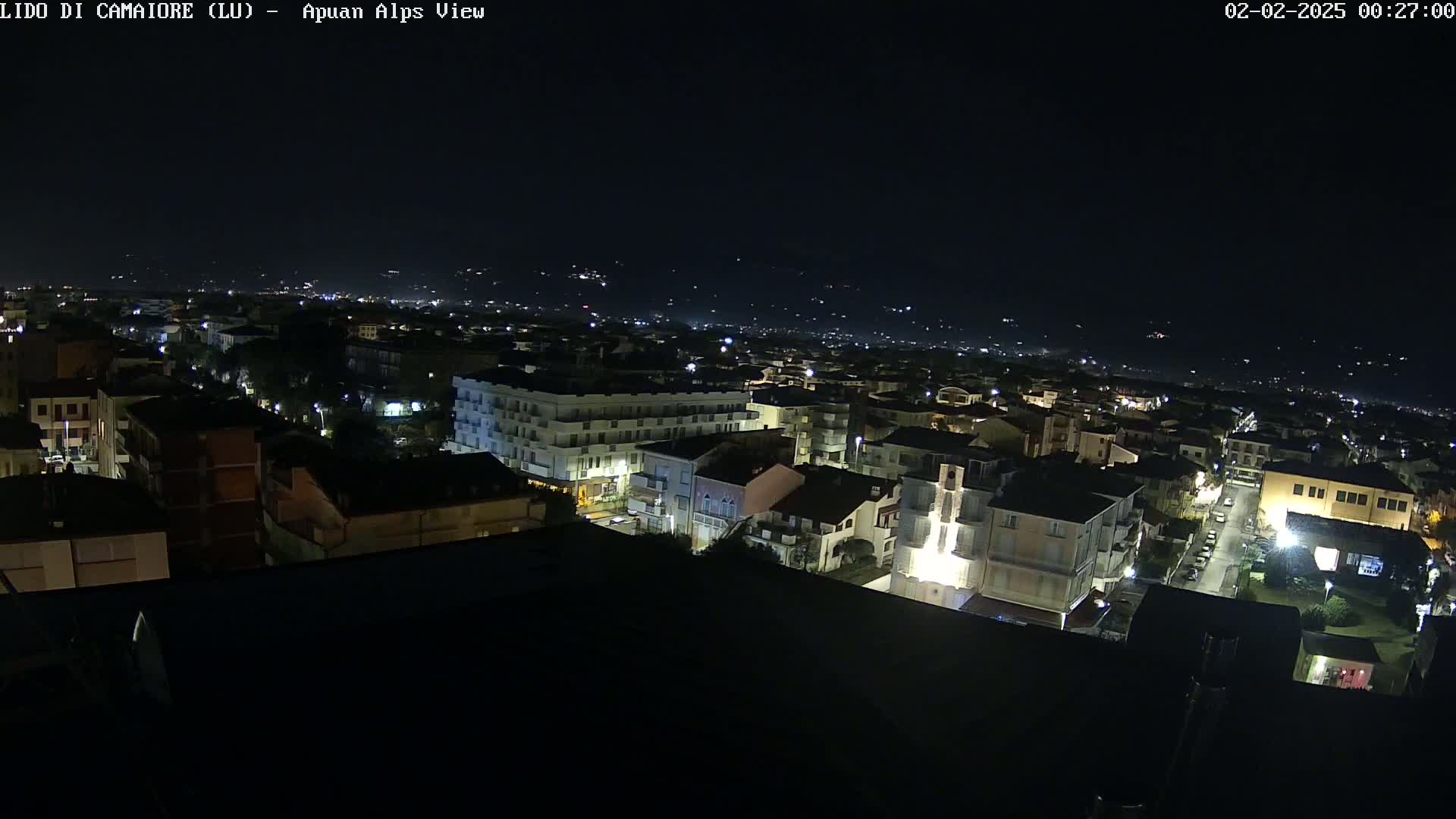 A nighttime aerial view of a town, illuminated by streetlights, with distant hills visible in the background under a clear, dark sky.