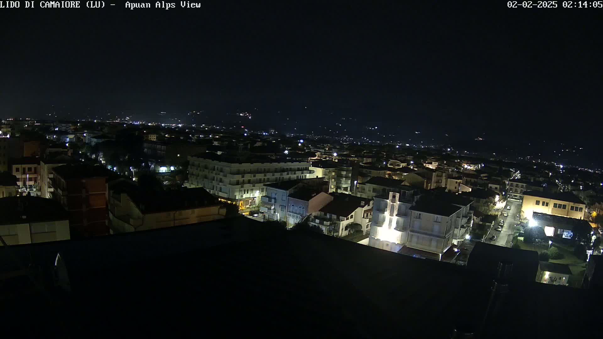 A nighttime, high-angle view of a city illuminated by streetlights, with distant hills faintly visible in the background under a dark sky.