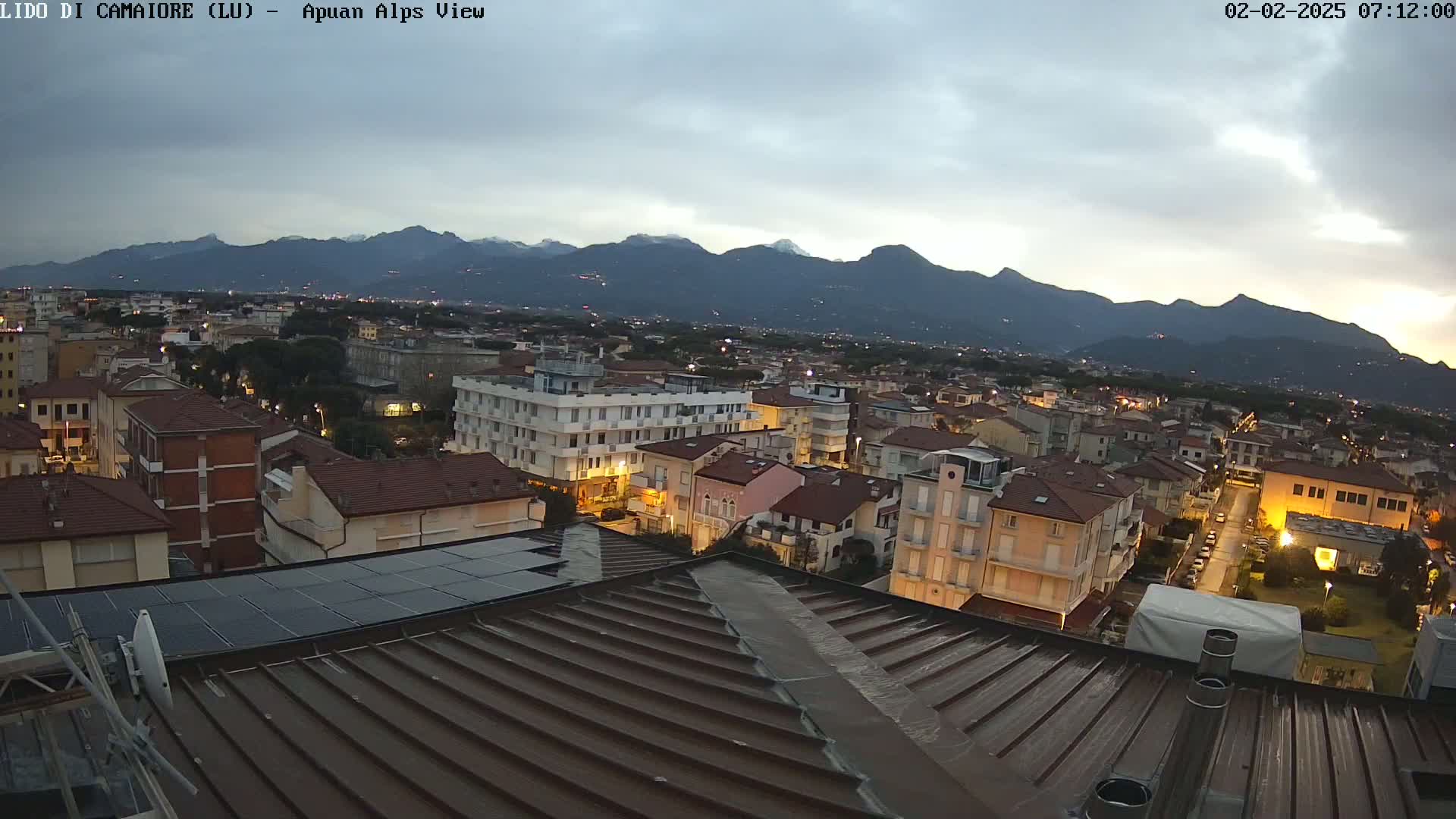 A view from a rooftop shows a town at dusk, with mountains in the background under a cloudy sky.