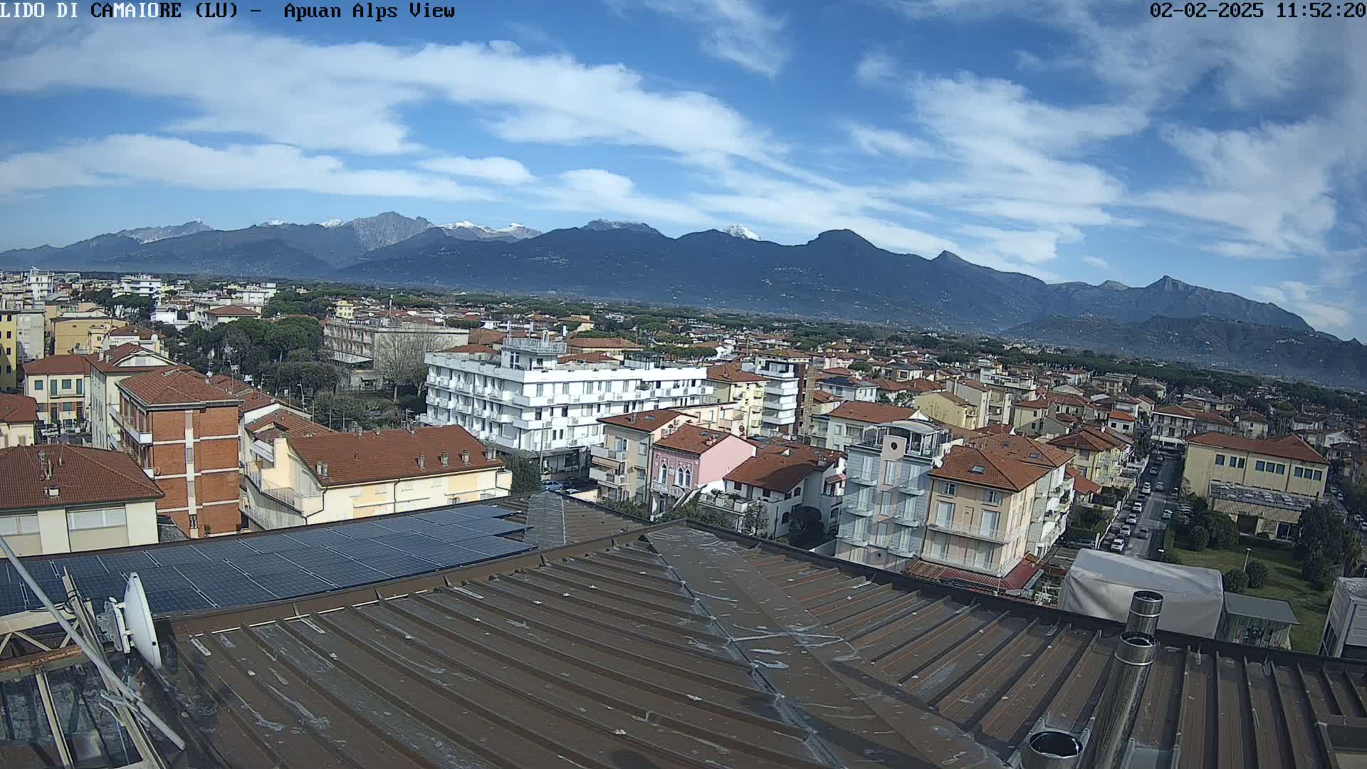 A partly cloudy blue sky overlooks a town of low-rise buildings with red-tiled roofs,  set against a backdrop of snow-capped mountains in the distance.