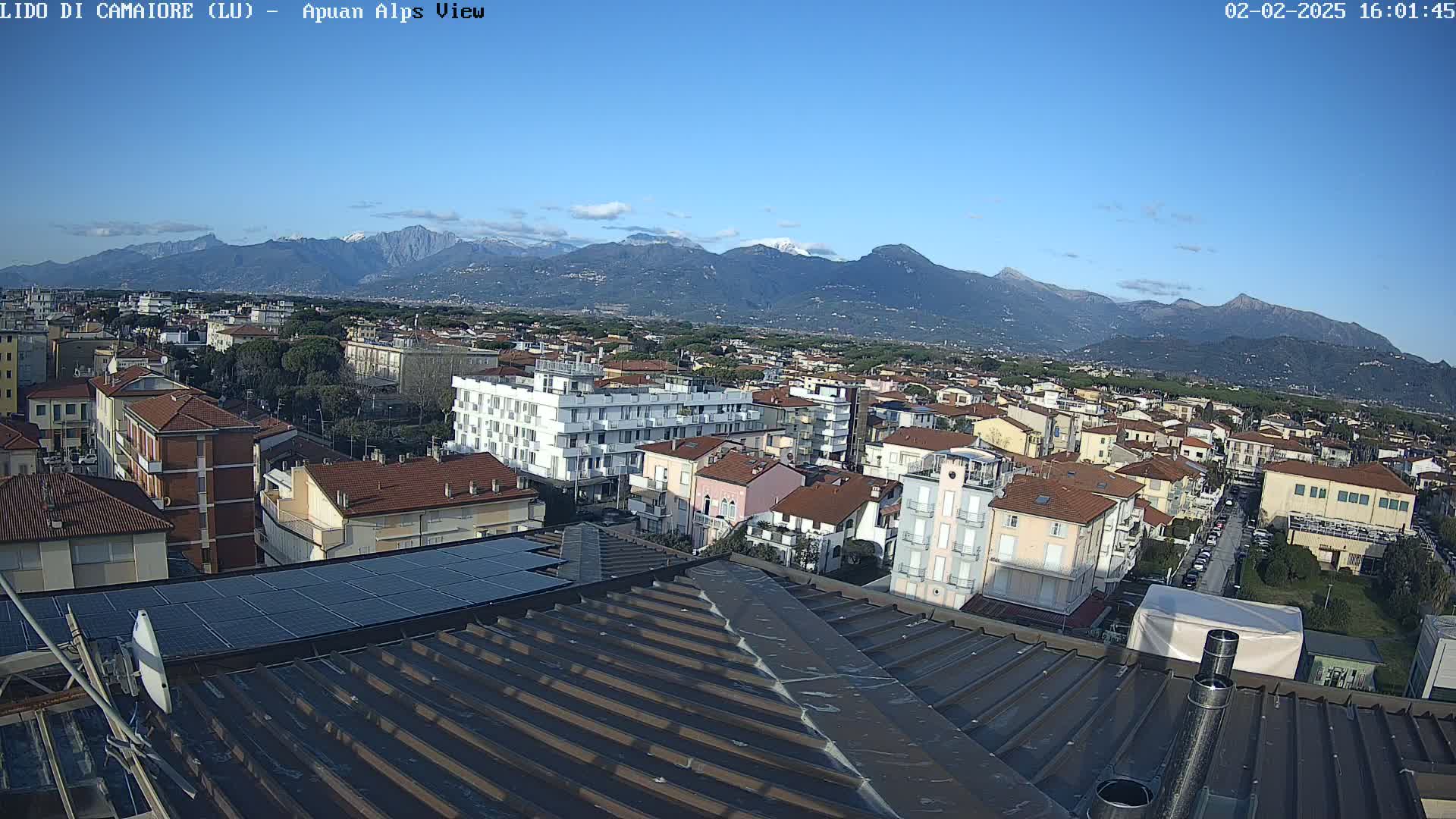 A clear, sunny day shows a view of a town nestled at the foot of snow-capped mountains, as seen from a rooftop with solar panels.
