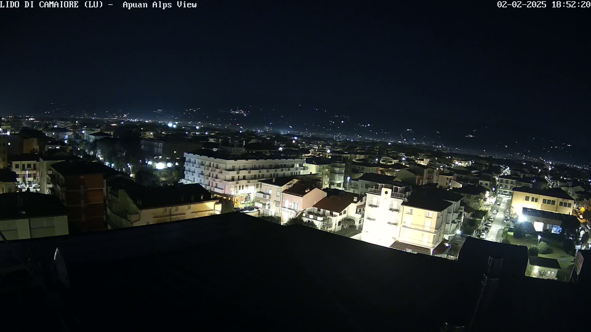 A nighttime aerial view of a town, illuminated by streetlights, with distant hills faintly visible under a clear, dark sky.