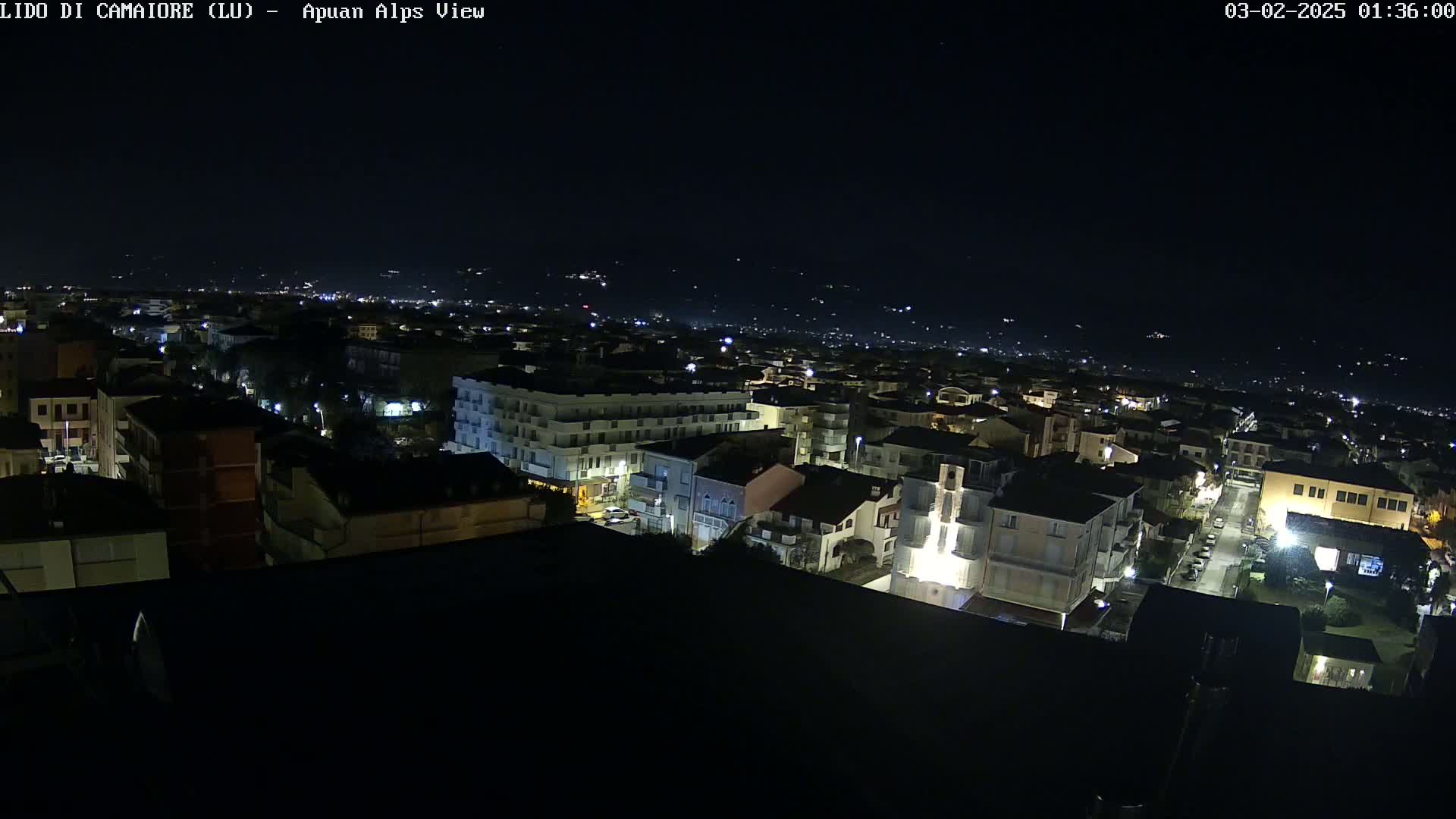 A nighttime, aerial view of a town illuminated by streetlights, with distant hills faintly visible under a clear, dark sky.
