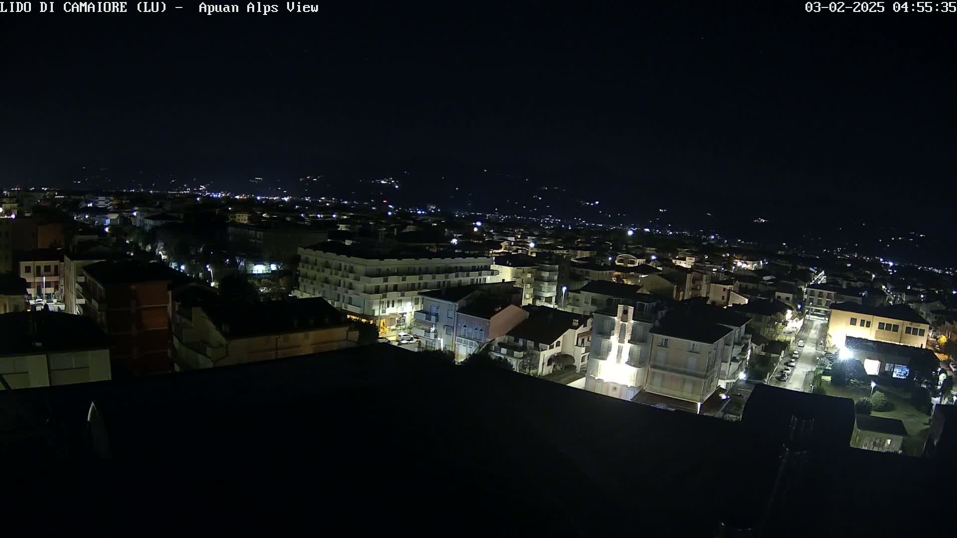 A nighttime aerial view of a town illuminated by streetlights, with a dark, clear sky and distant hills visible in the background.