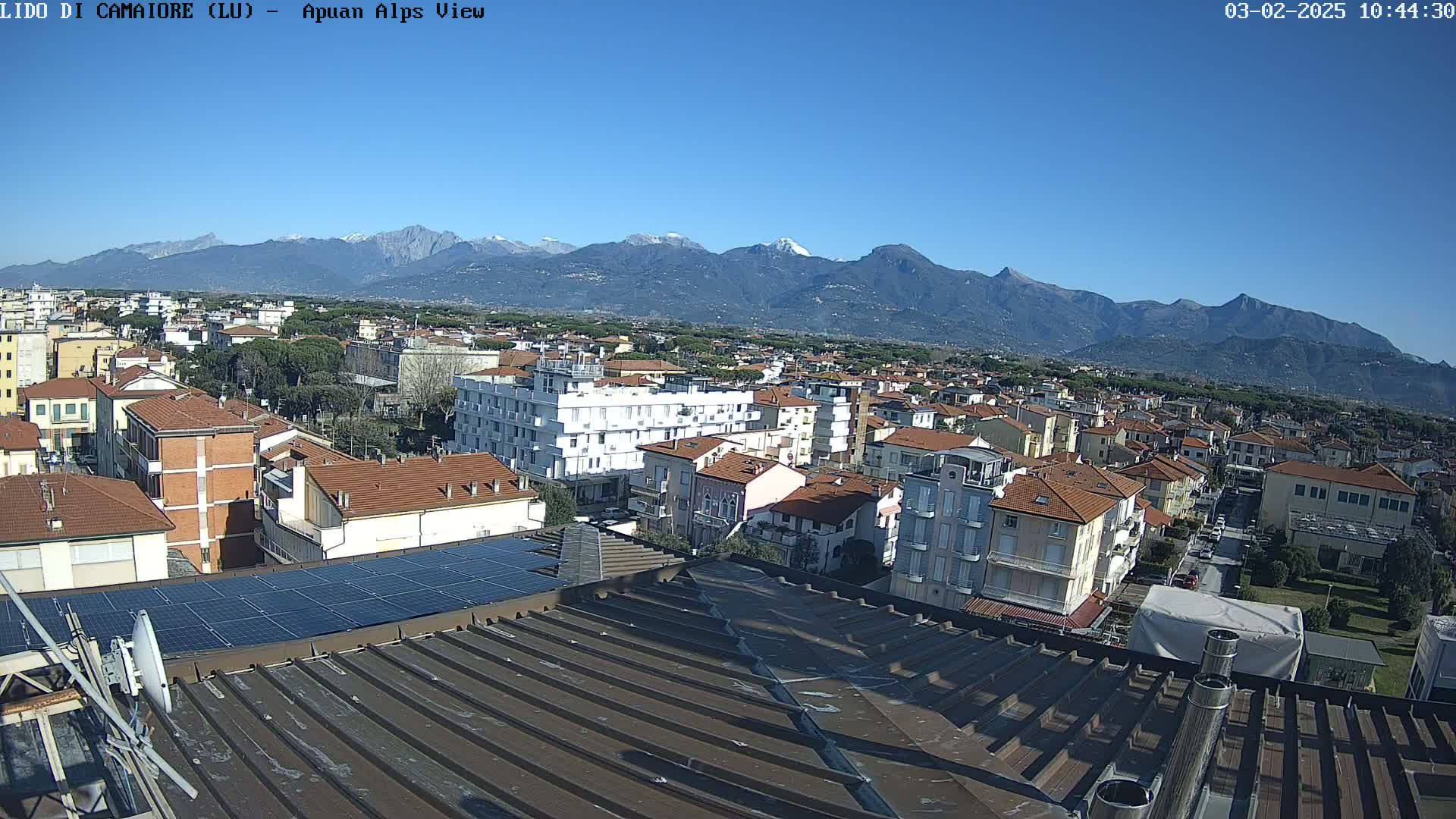 A rooftop view of a town shows many buildings under a clear blue sky, with snow-capped mountains in the distance.