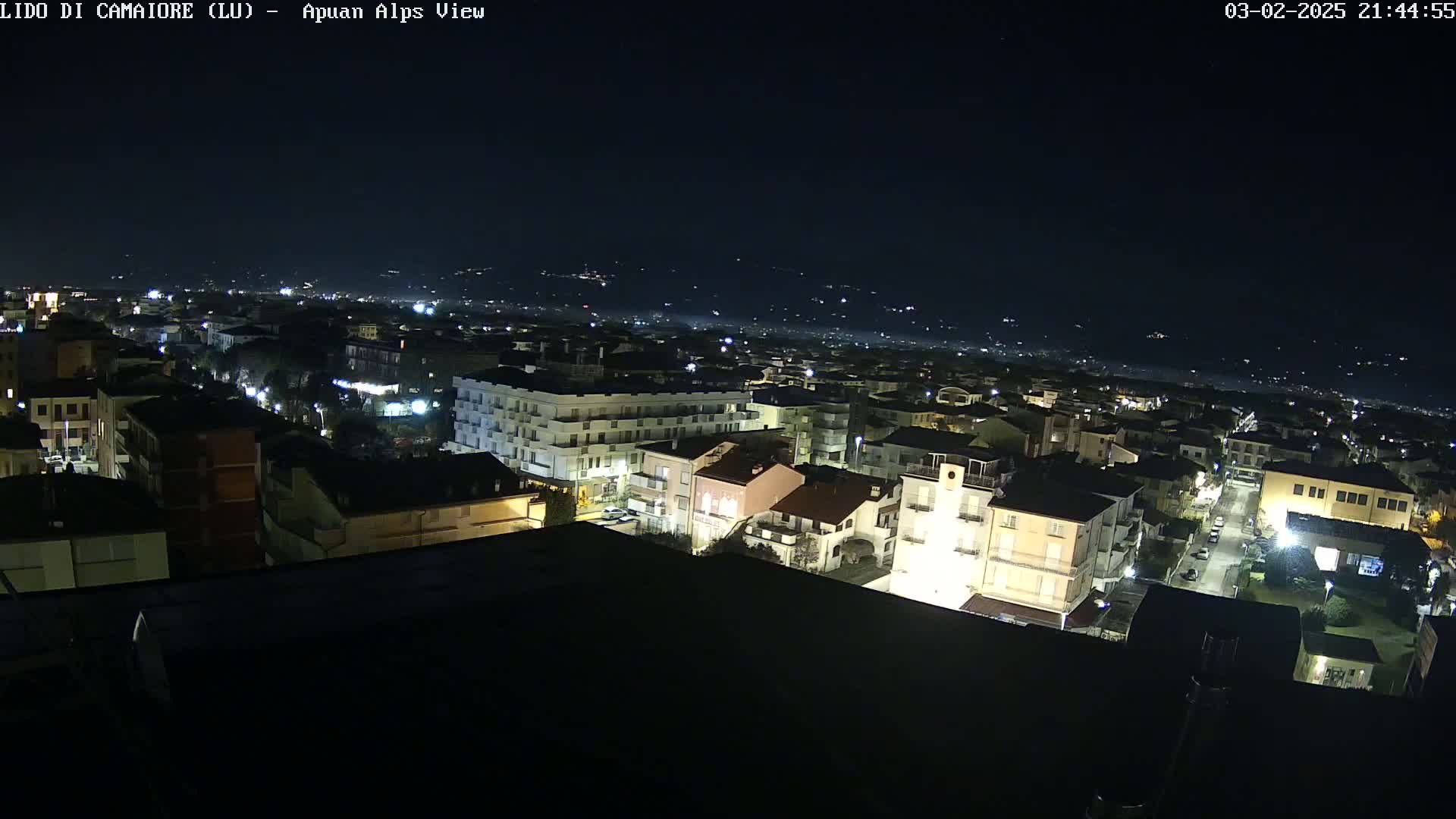 A nighttime aerial view of a town illuminated by streetlights, with a dark, clear sky and distant hills visible in the background.