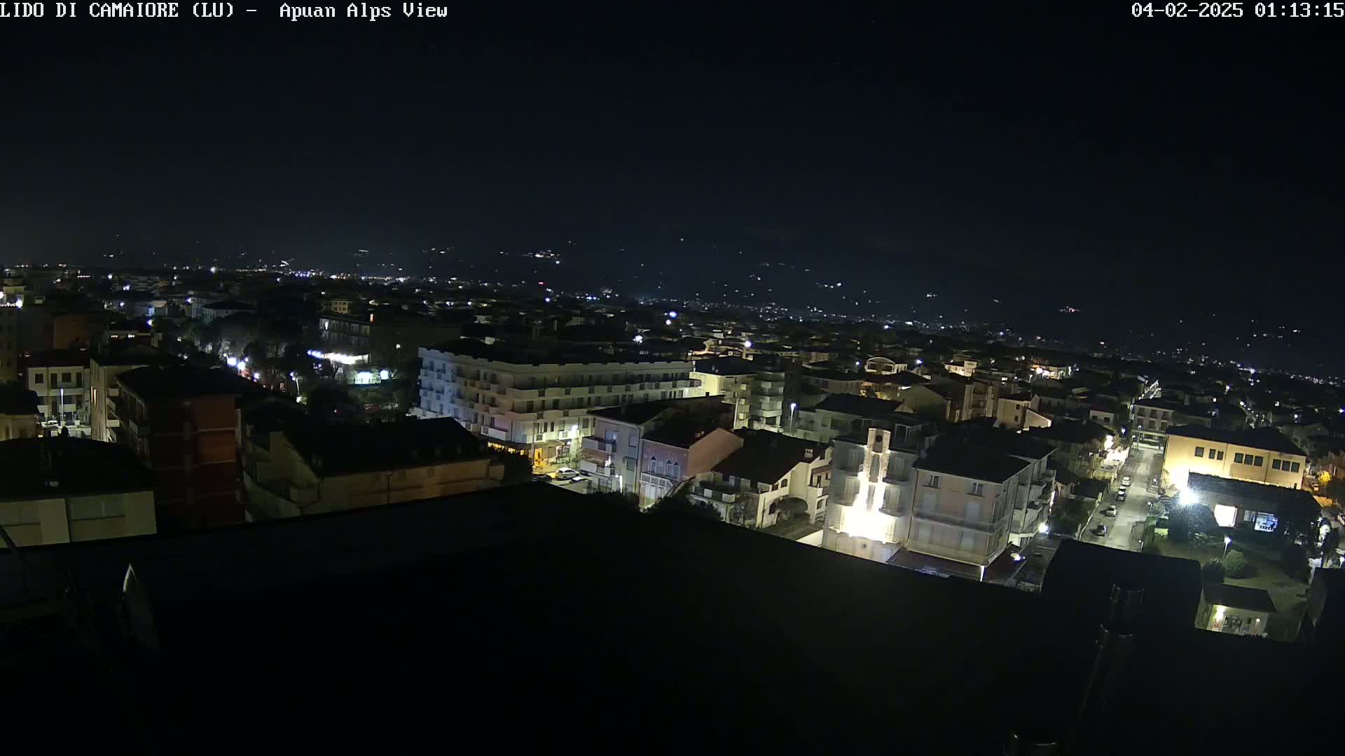 A nighttime aerial view of a town illuminated by streetlights, with a dark sky and distant lights visible in the background.