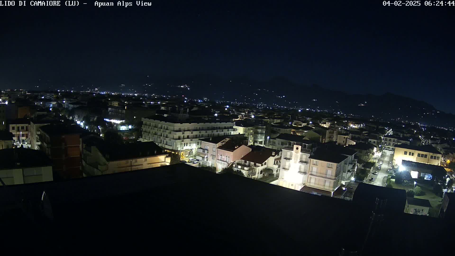 A nighttime aerial view shows a city illuminated by streetlights, with dark mountains visible in the distance under a clear, dark sky.