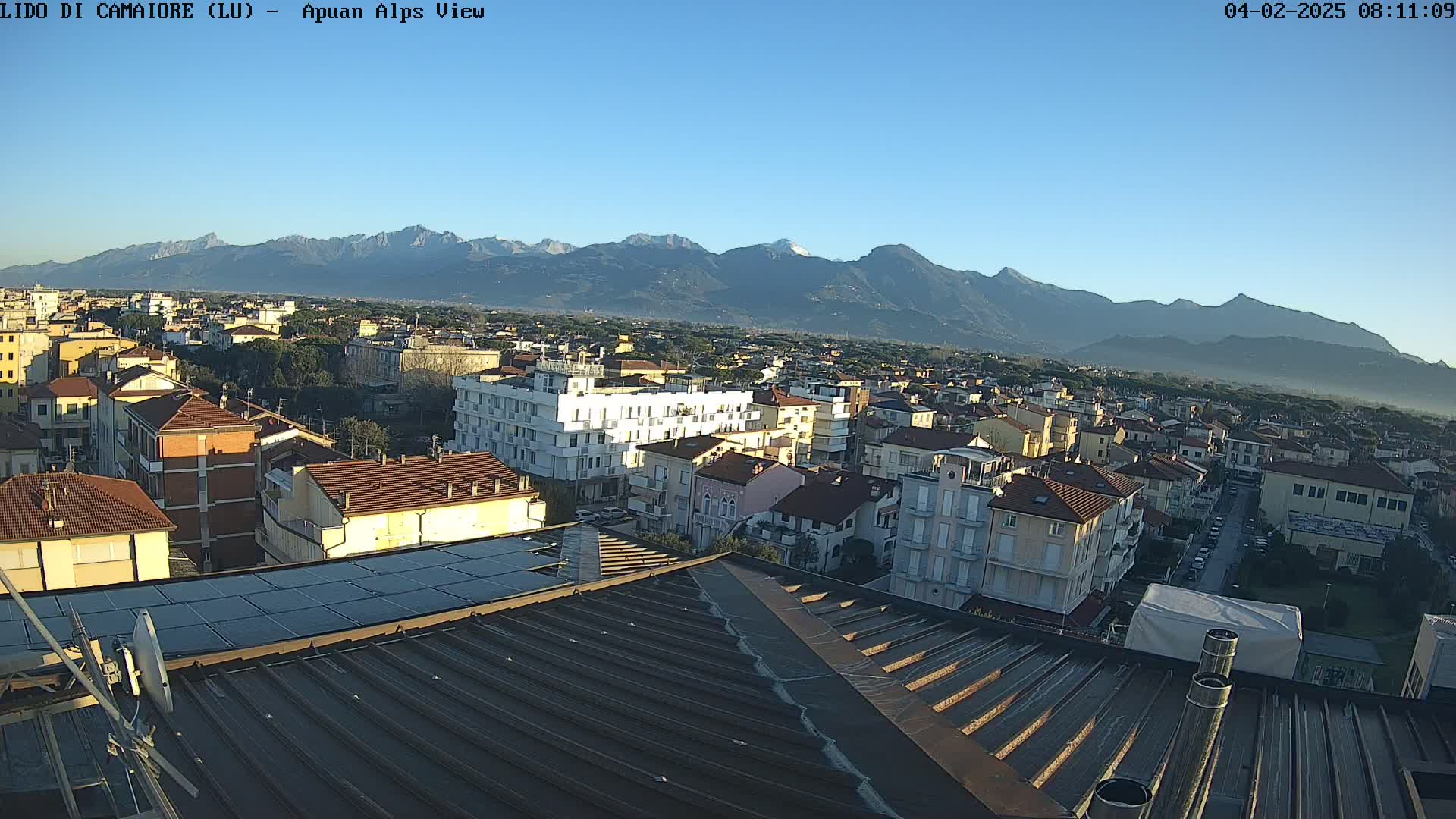 A rooftop view of a town nestled at the foot of a mountain range under a clear, sunny sky.
