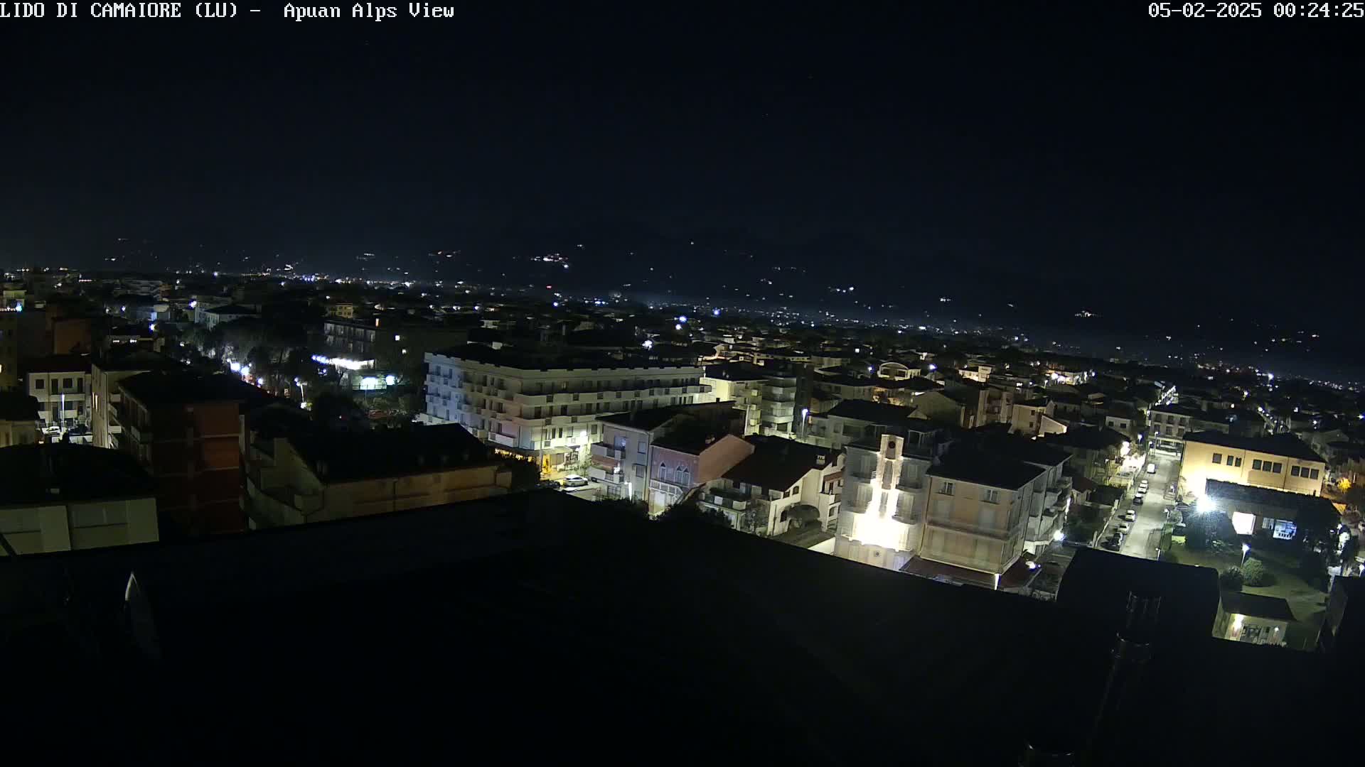 A nighttime aerial view of a town, showing illuminated buildings and streets, with a dark, clear sky and distant hills in the background.