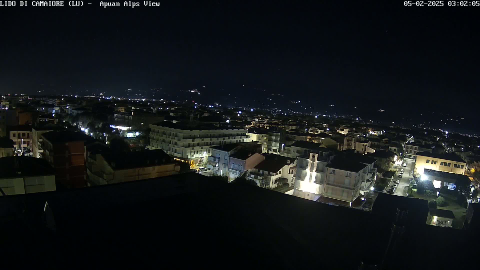 A nighttime, aerial view of a town with dimly lit buildings and streets, extending to a dark, distant mountain range under a clear sky.