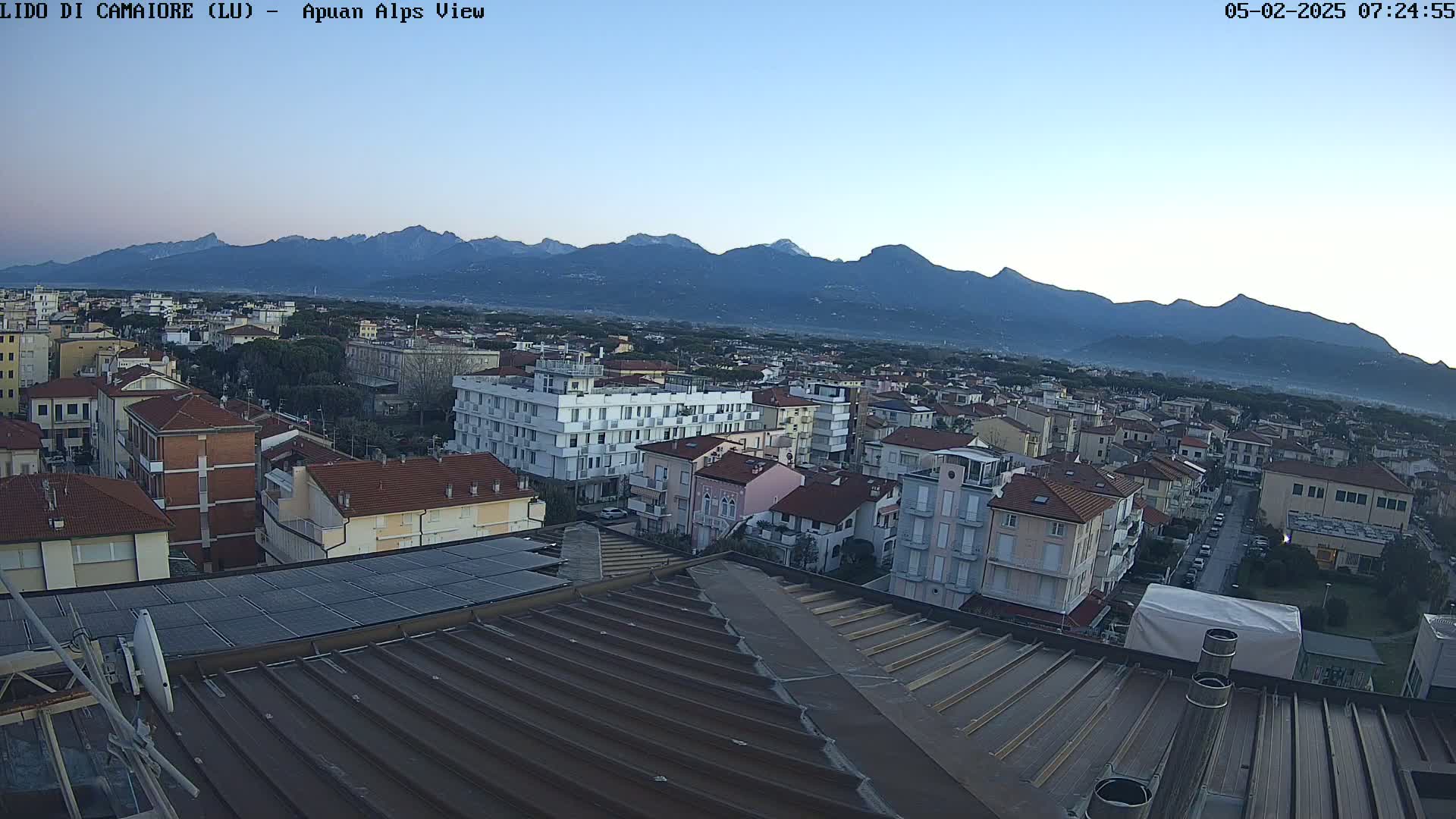 A view from a rooftop shows a town nestled below a mountain range under a clear, light-blue sky.