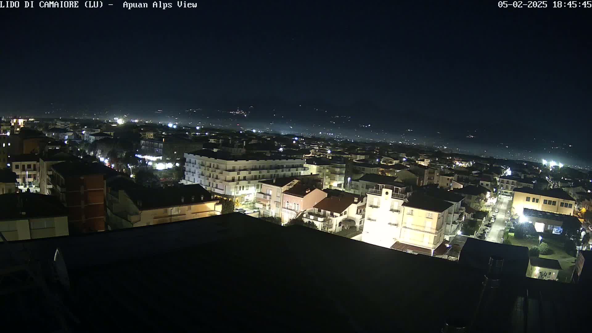 A nighttime aerial view shows a town illuminated by streetlights, with a hazy, dark sky and distant hills visible in the background.