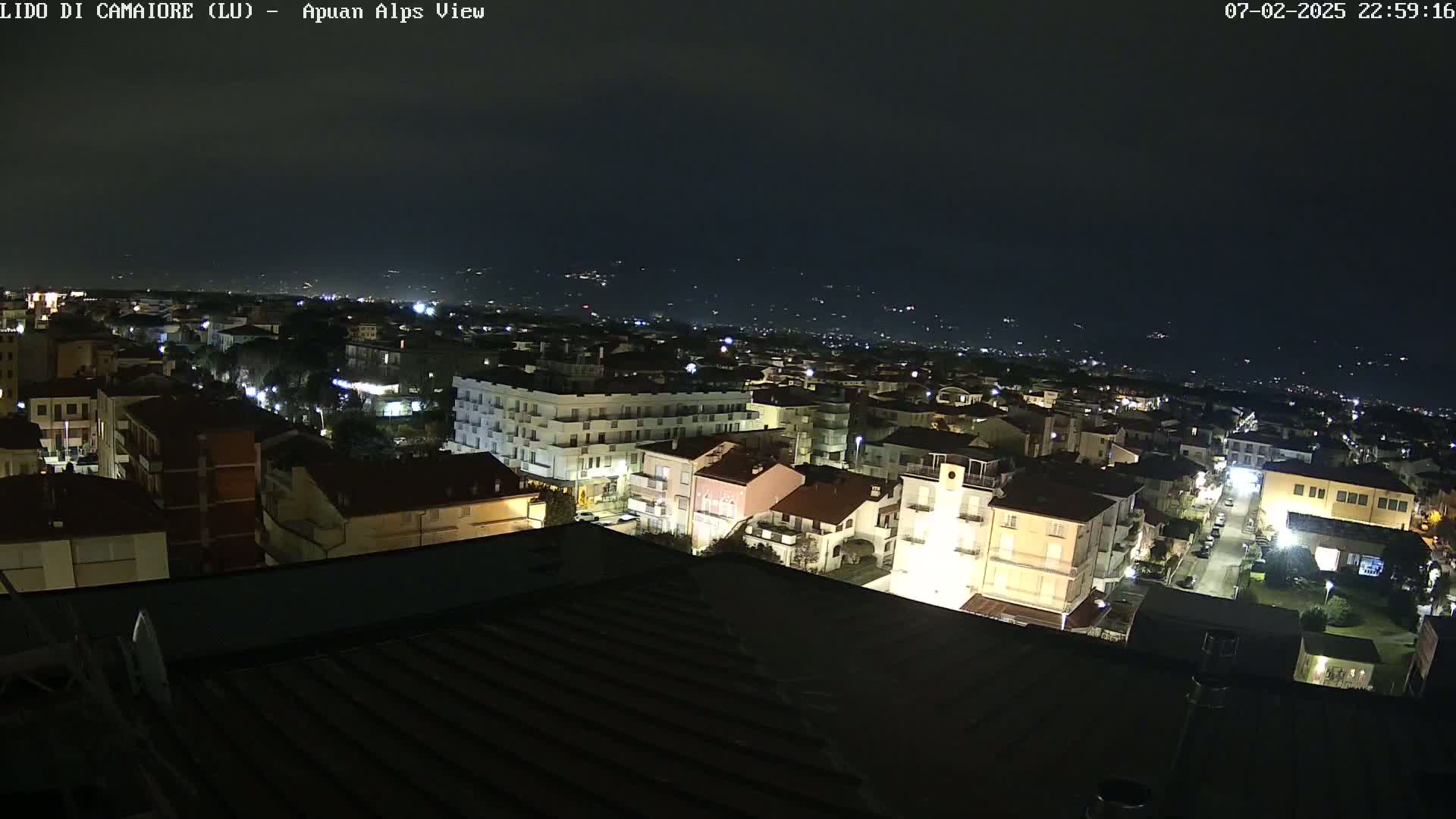 A nighttime, aerial view of a town illuminated by streetlights, with a dark sky and distant hills visible in the background.