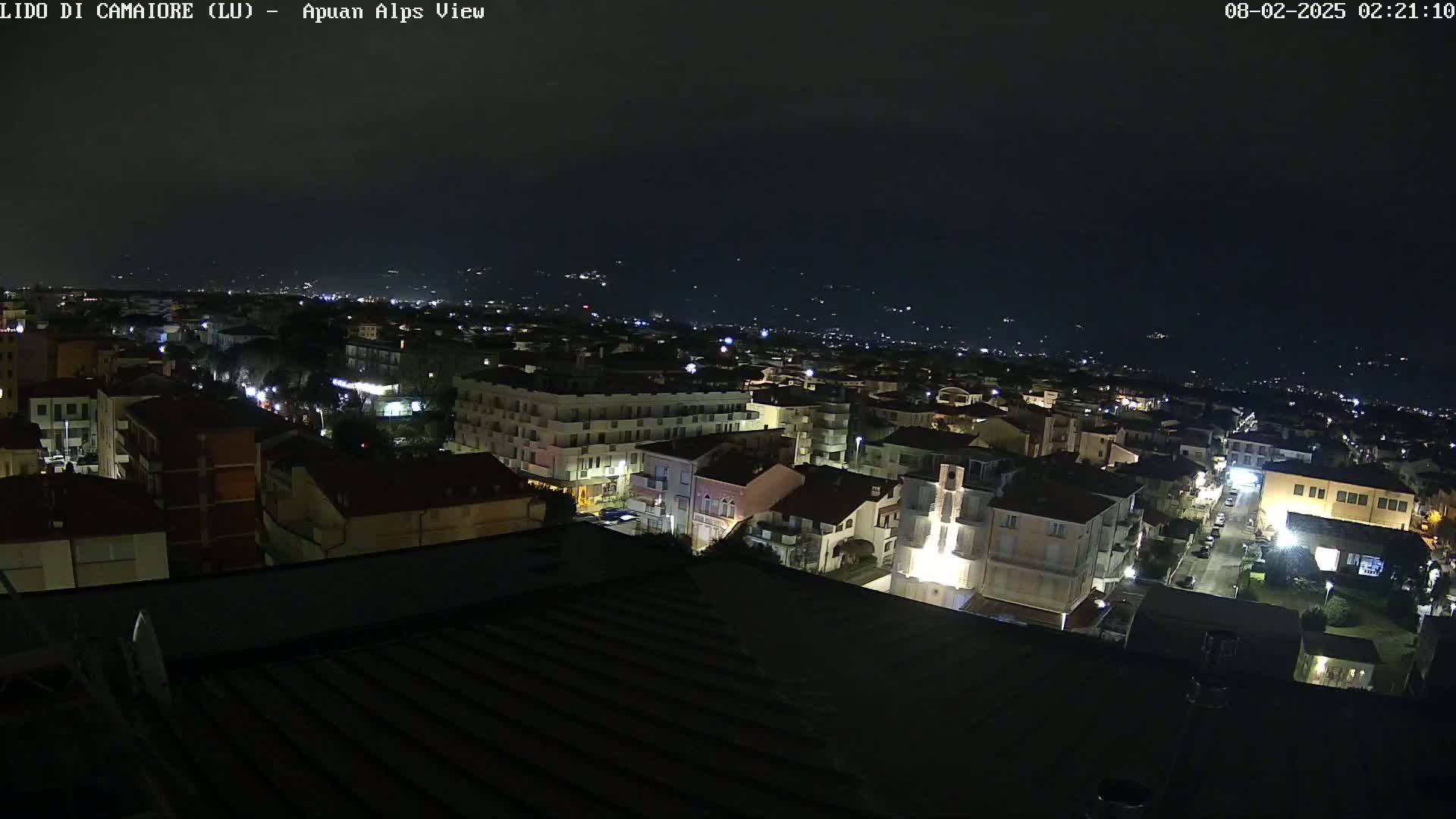 A nighttime aerial view of a town shows a cityscape illuminated by streetlights and building lights, with distant, dimly lit mountains in the background under a clear, dark sky.
