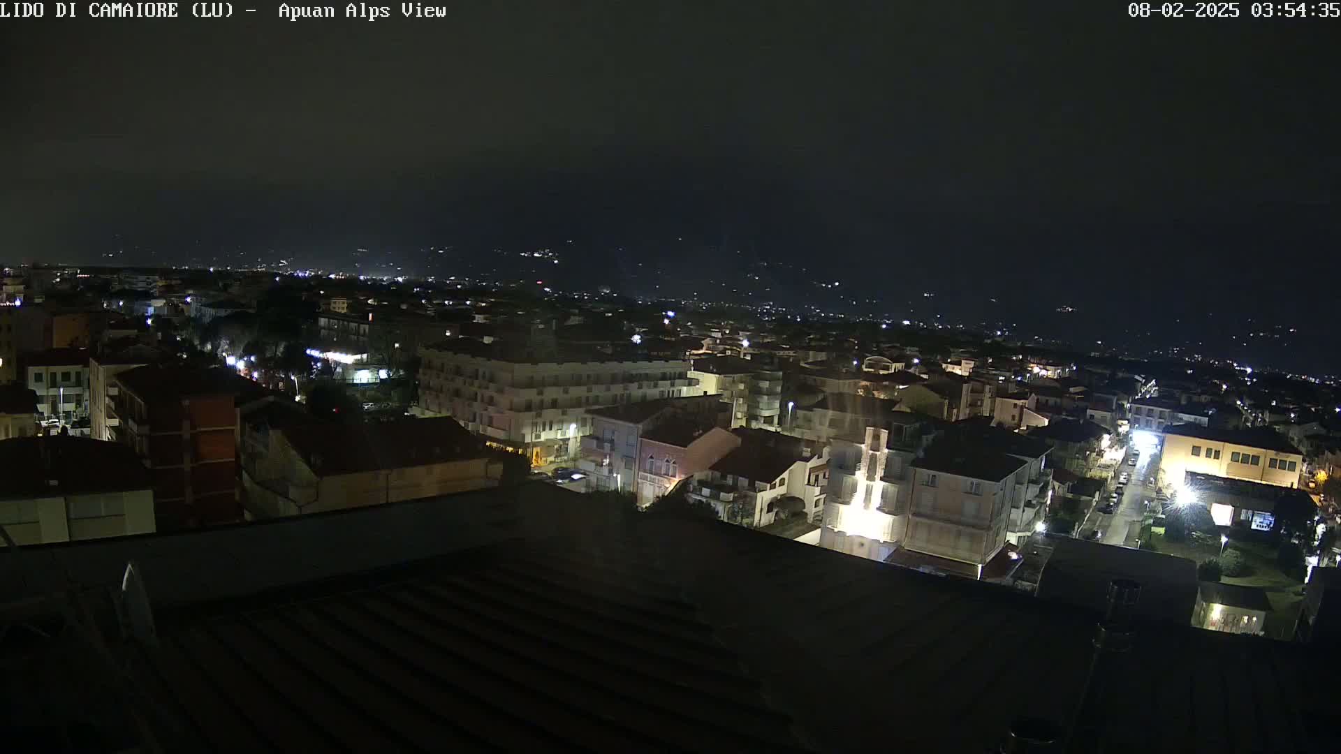 A nighttime aerial view of a town illuminated by streetlights, with distant hills faintly visible under a clear, dark sky.