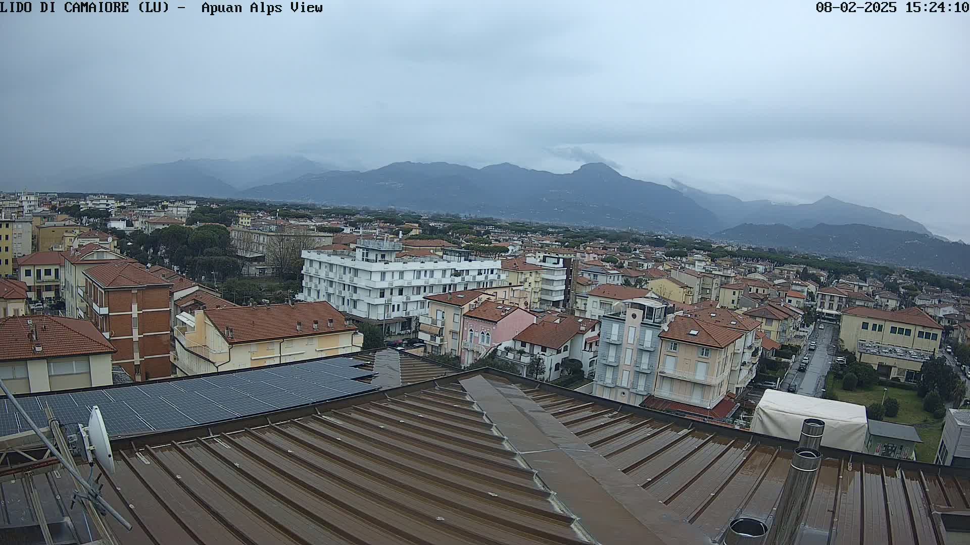 A wet, brown rooftop overlooks a town nestled against a range of mountains under an overcast sky.