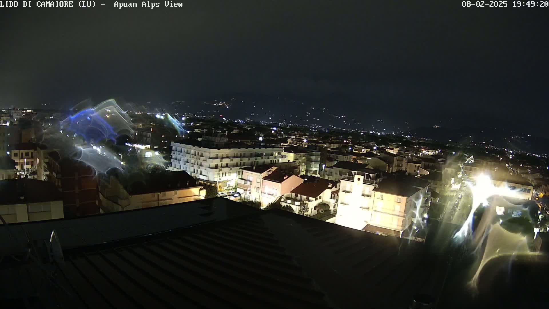 A nighttime view of a town illuminated by streetlights, with a blurry effect possibly due to rain or camera distortion, overlooking distant hills.