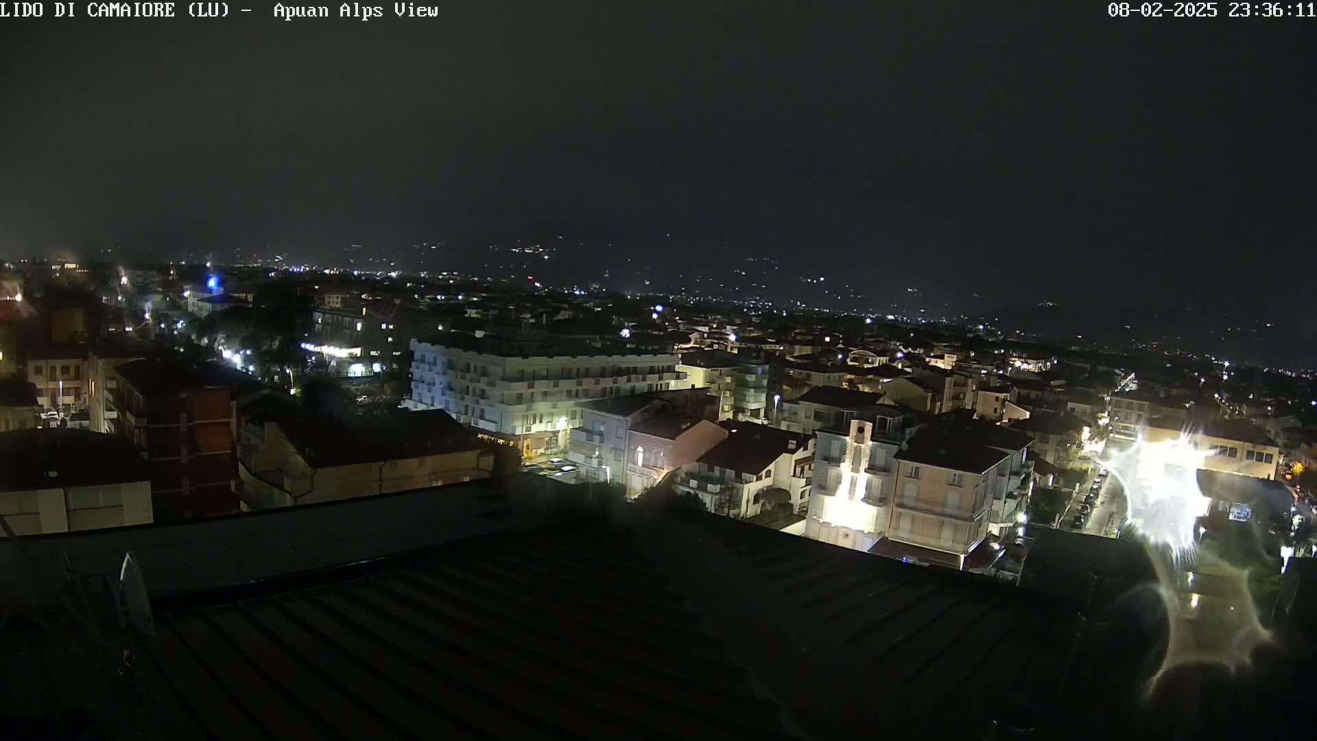 A nighttime view of a town illuminated by streetlights, with distant hills faintly visible under a dark sky.