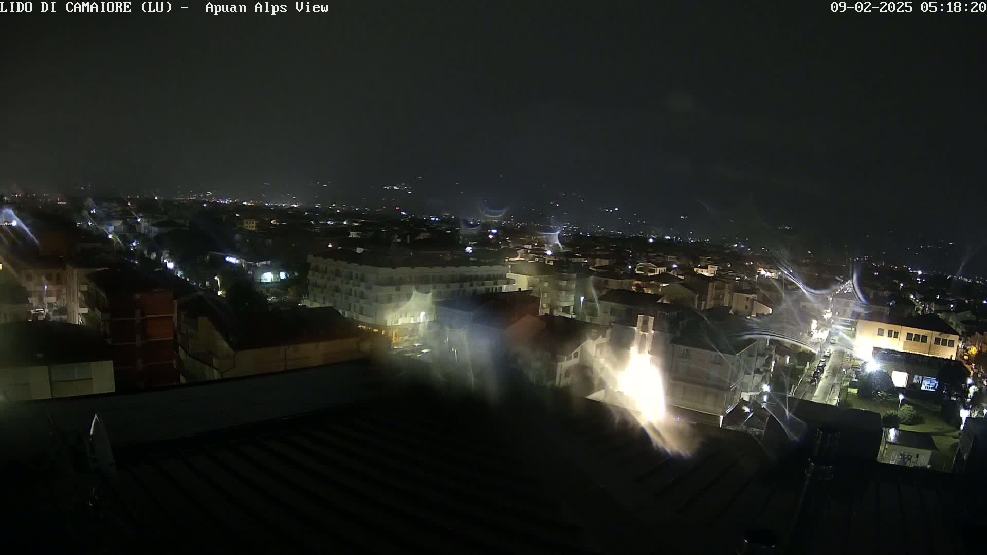 A nighttime aerial view of a town shows rain-streaked lights on buildings and streets under a dark sky.