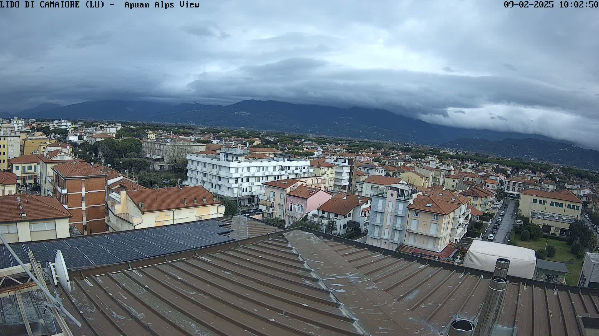 A view from a rooftop shows a town under a cloudy sky with mountains in the distance.