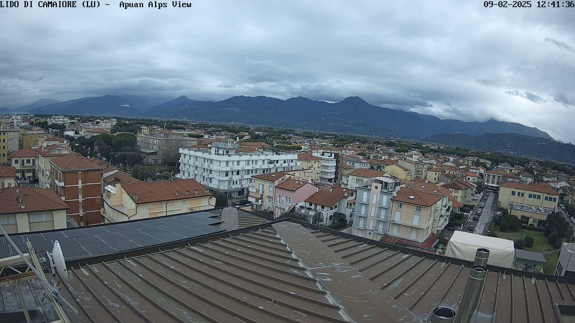 A view from a rooftop shows a town nestled against a mountain range under an overcast sky.