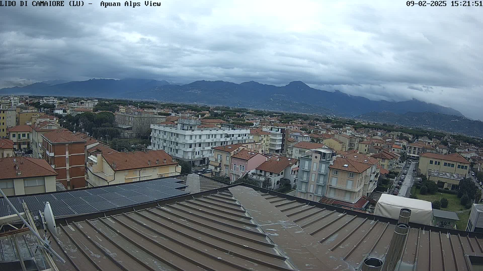 A view from a rooftop shows a town under a cloudy sky with distant mountains.