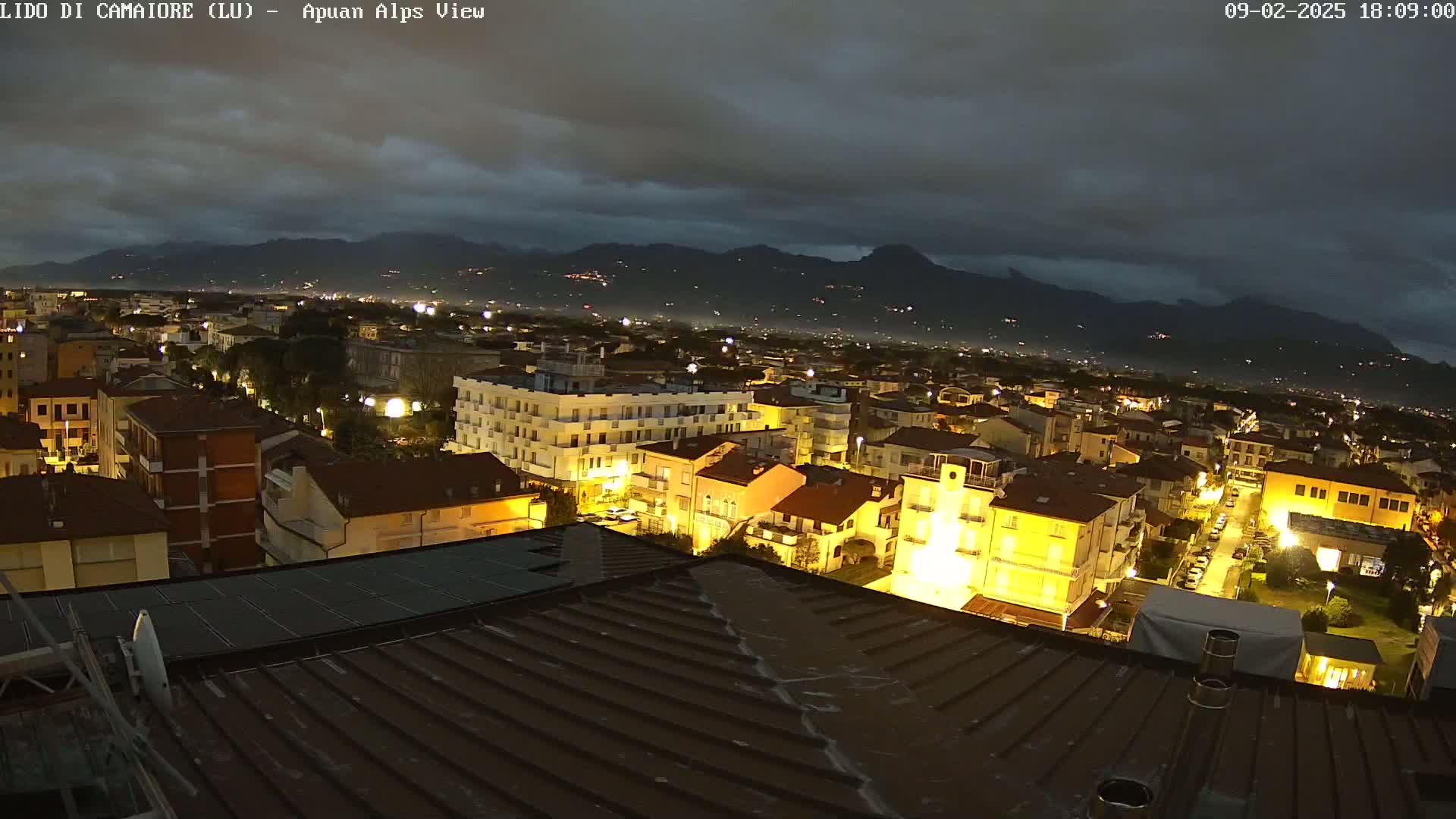 A nighttime view of a town illuminated by streetlights, situated below dark, cloudy skies and a mountain range in the distance.