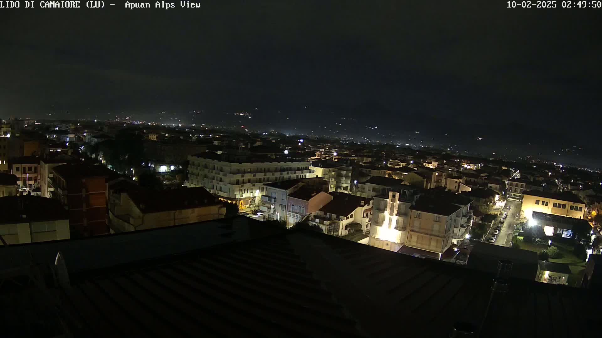 A nighttime aerial view of a town, illuminated by streetlights, with a distant mountain range visible under a dark, clear sky.