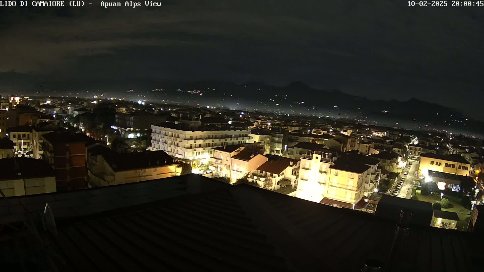 A nighttime aerial view of a town illuminated by streetlights, with a mountain range faintly visible in the background under a dark, cloudy sky.