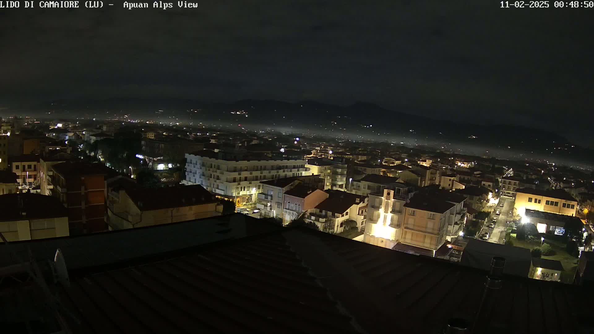 A nighttime aerial view of a town illuminated with streetlights, nestled beneath a dark, hazy mountain range.