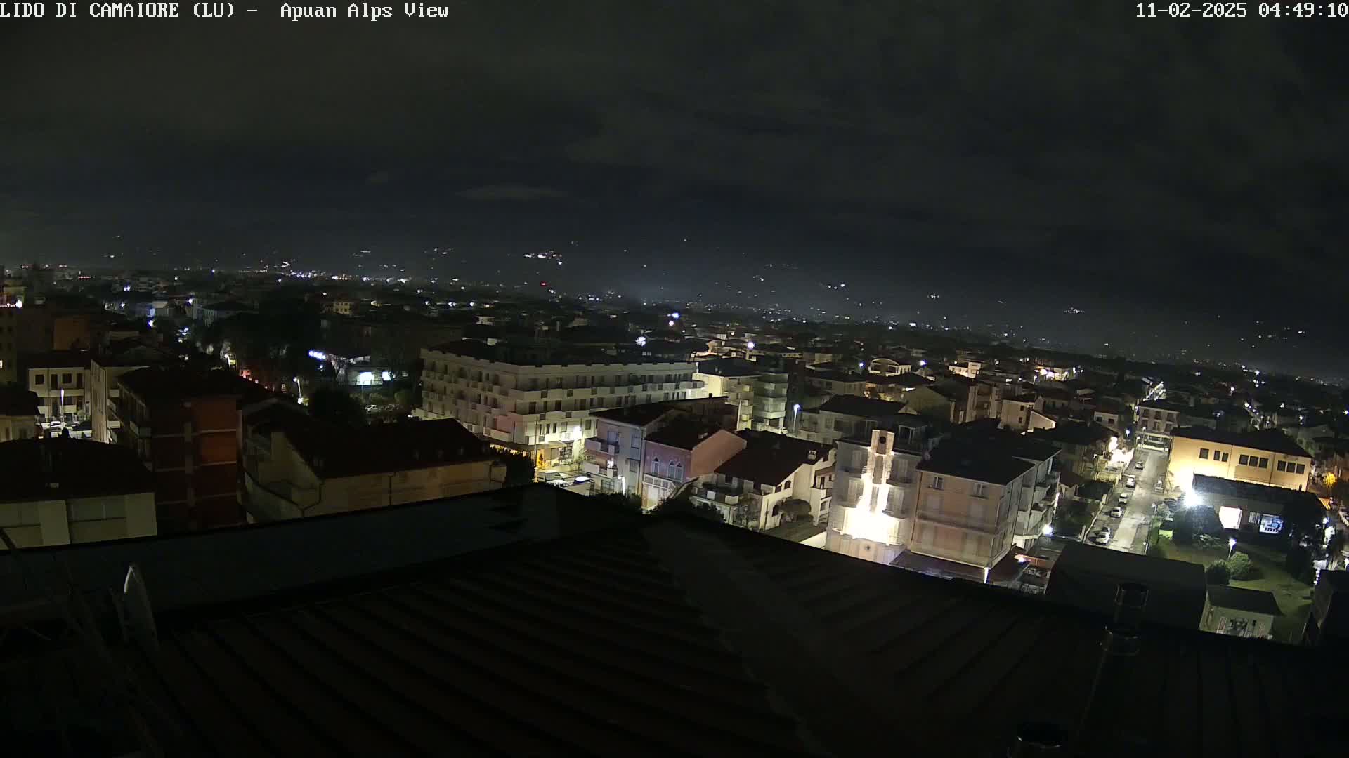 A nighttime aerial view of a town shows many buildings illuminated, under a partly cloudy sky.