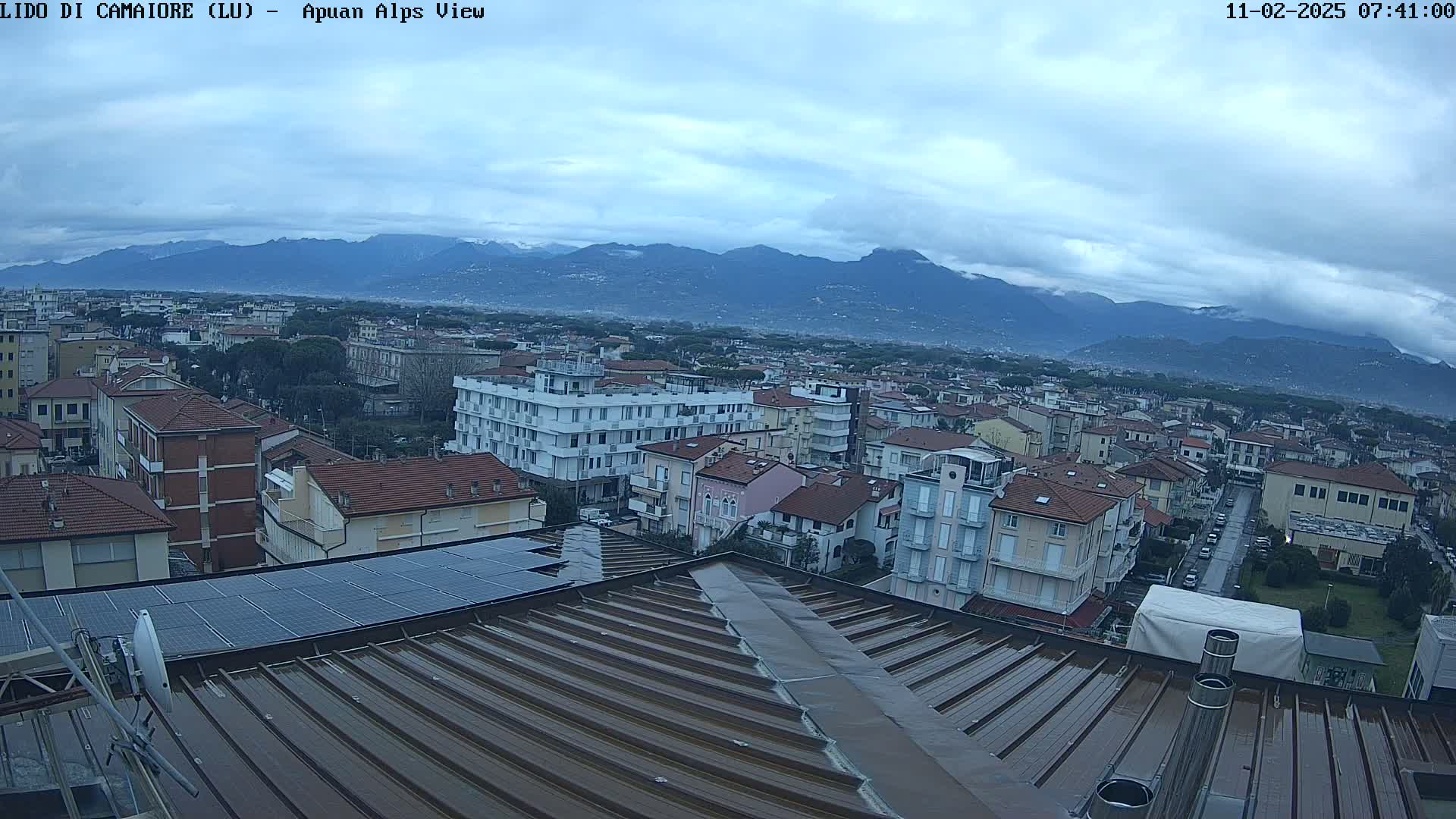 A view of a town under a cloudy sky, with mountains in the background and a wet rooftop in the foreground.