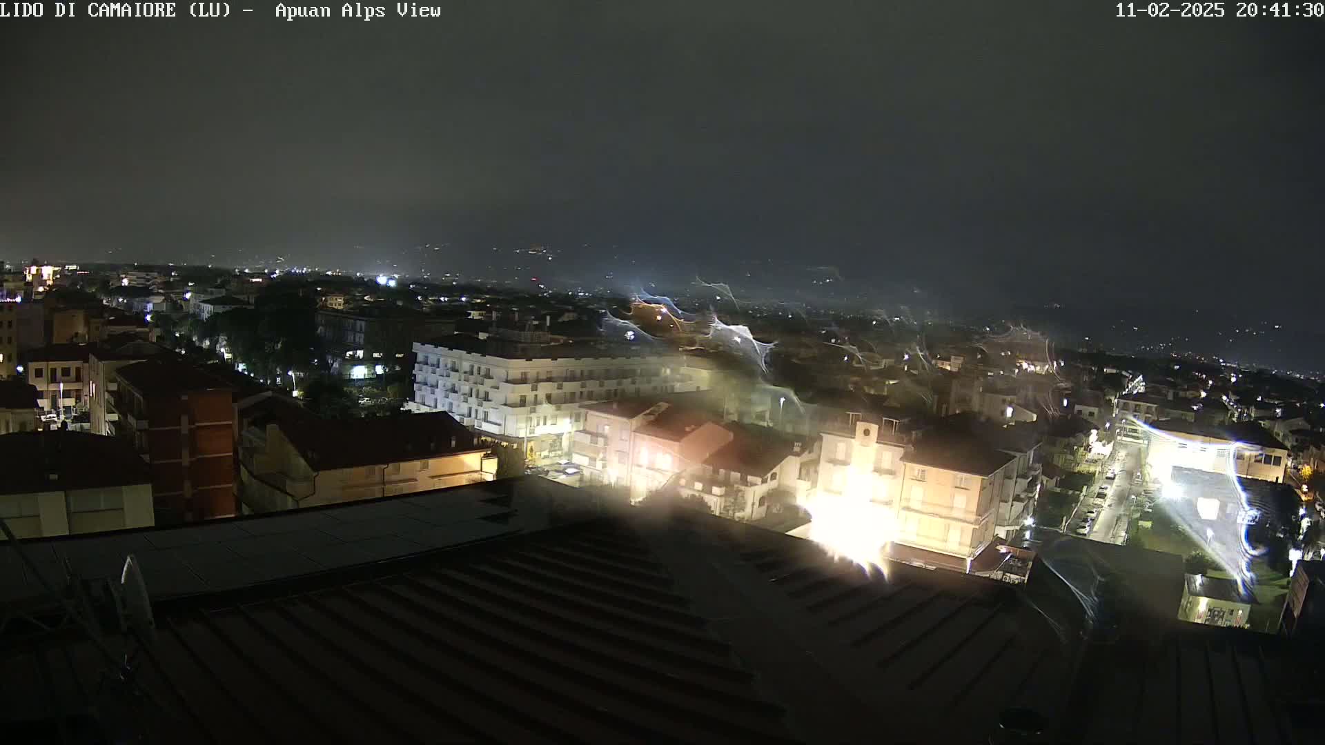 A nighttime aerial view of a city, illuminated by streetlights, with streaks of light possibly from rain or wind in the dark sky.