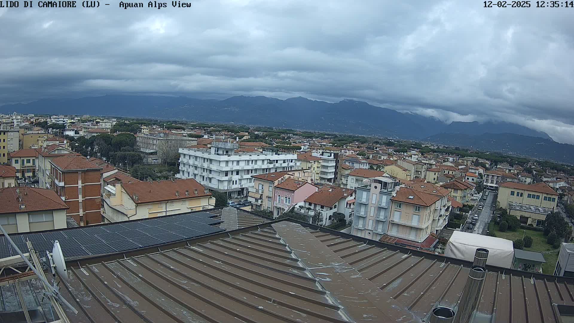 A view from a rooftop shows a town of low-rise buildings under a cloudy sky with mountains in the distance.