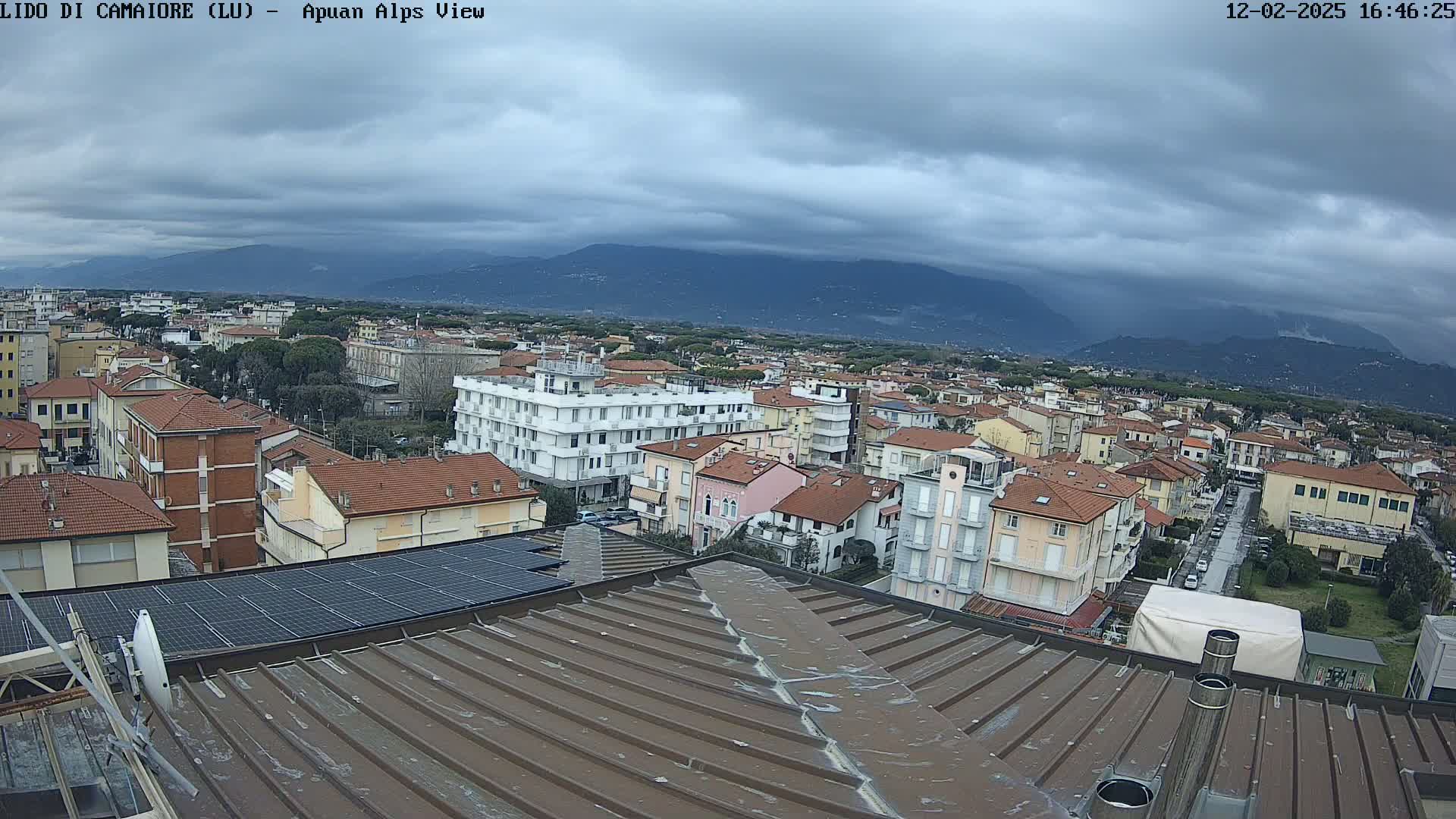 A view from a rooftop shows a town with many low-rise buildings under a cloudy sky and distant mountains.
