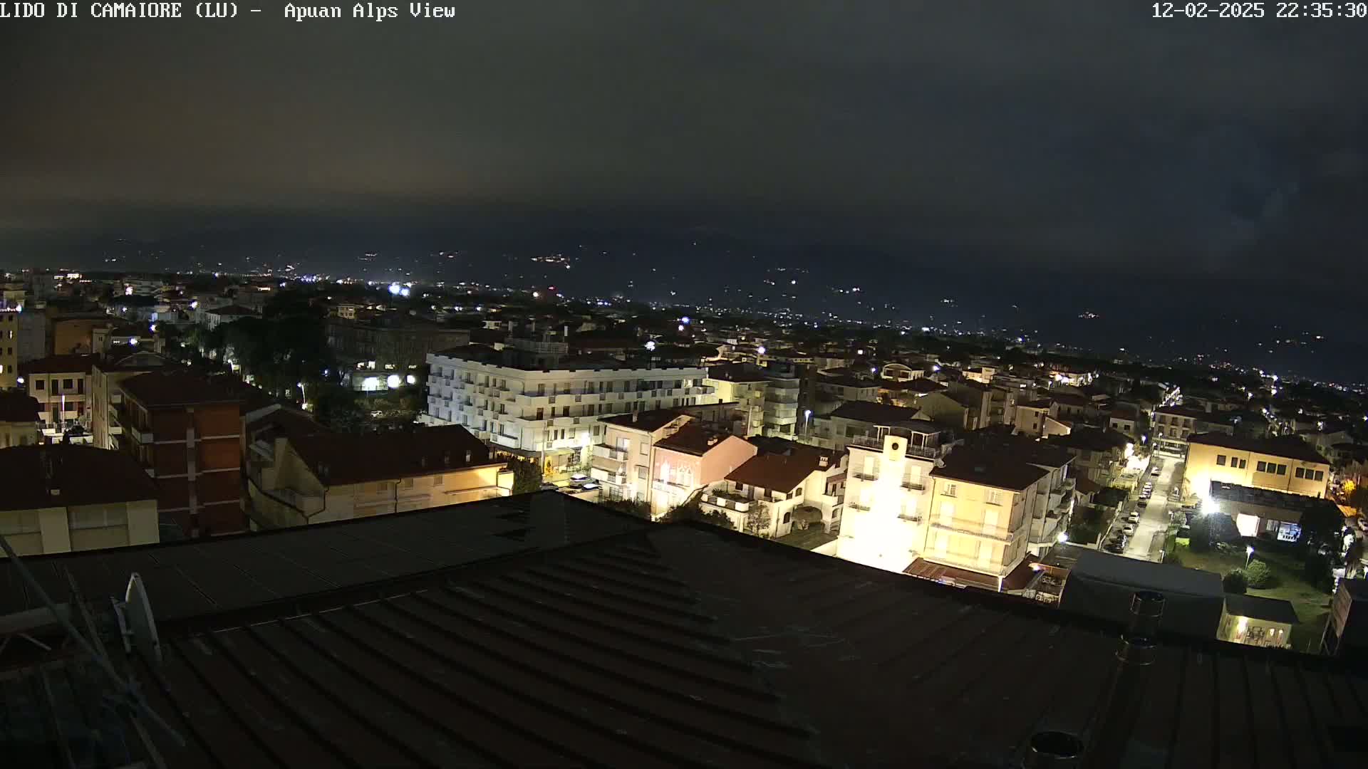 A nighttime, aerial view of a town illuminated by streetlights, with dark, cloudy skies and distant, dimly lit mountains in the background.