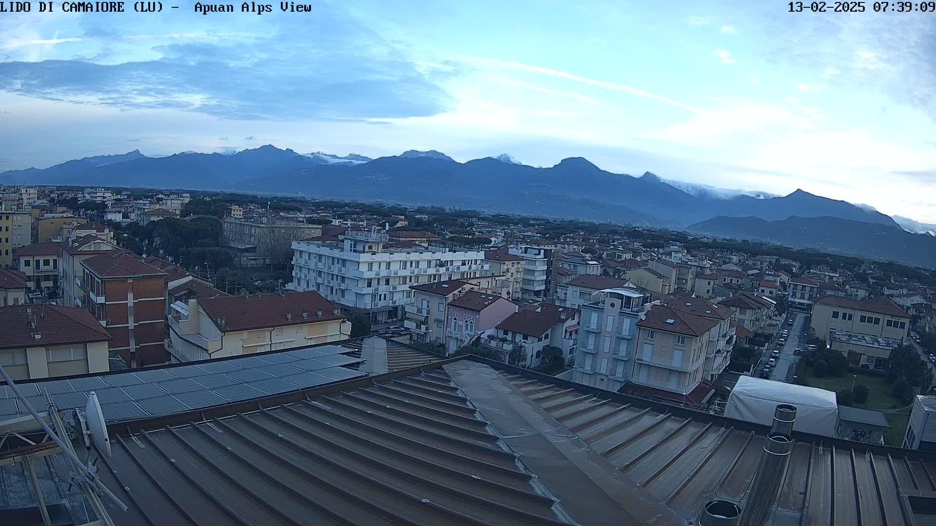 A rooftop view of a town nestled in a valley, overlooked by a range of mountains under a partly cloudy sky.