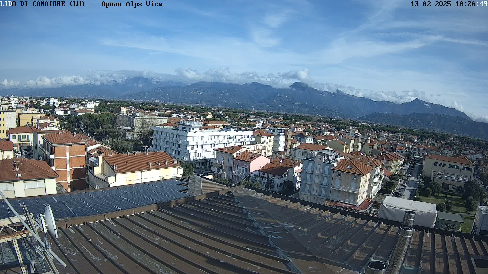 A rooftop view overlooking a town nestled beneath a mountain range on a partly cloudy day.