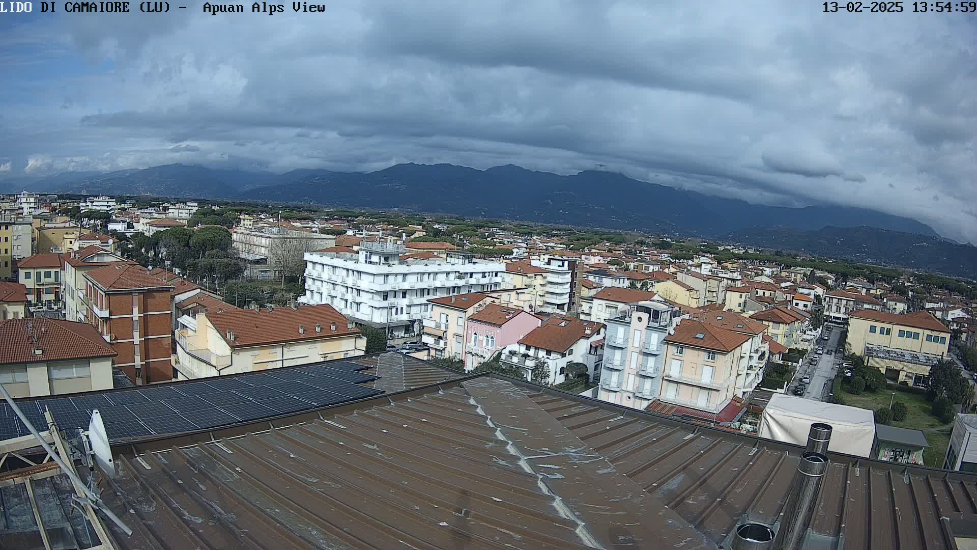 A view from a rooftop shows a town of low-rise buildings under a cloudy sky, with mountains in the distance.