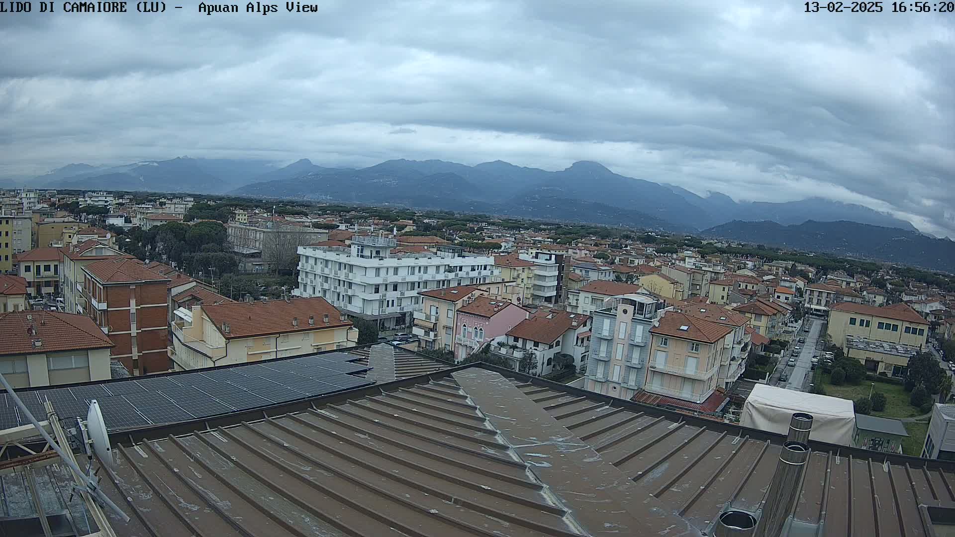 A view from a rooftop shows a town of low-rise buildings under a cloudy sky, with mountains in the distance.