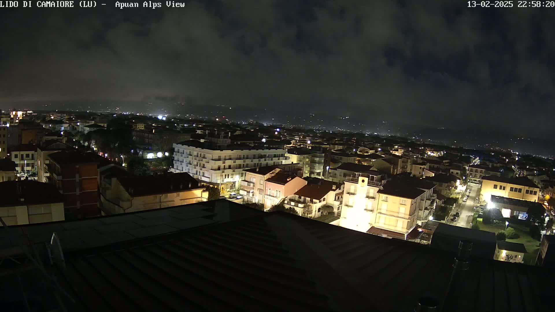 A nighttime, cloudy view of a town, with many buildings illuminated by streetlights and some distant lights visible in the background.