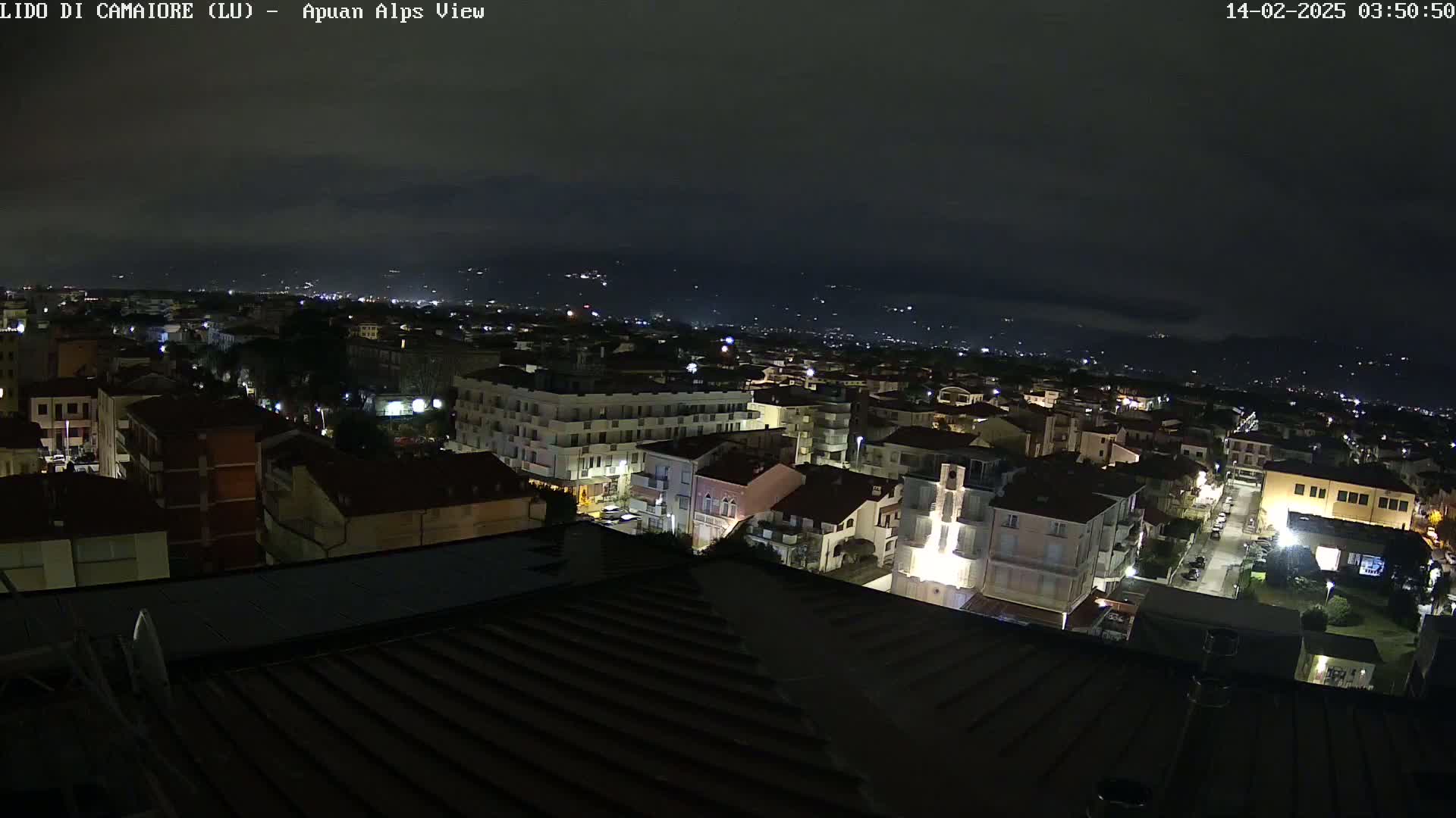 A nighttime view of a town illuminated by streetlights, with distant mountains visible under a dark, cloudy sky.