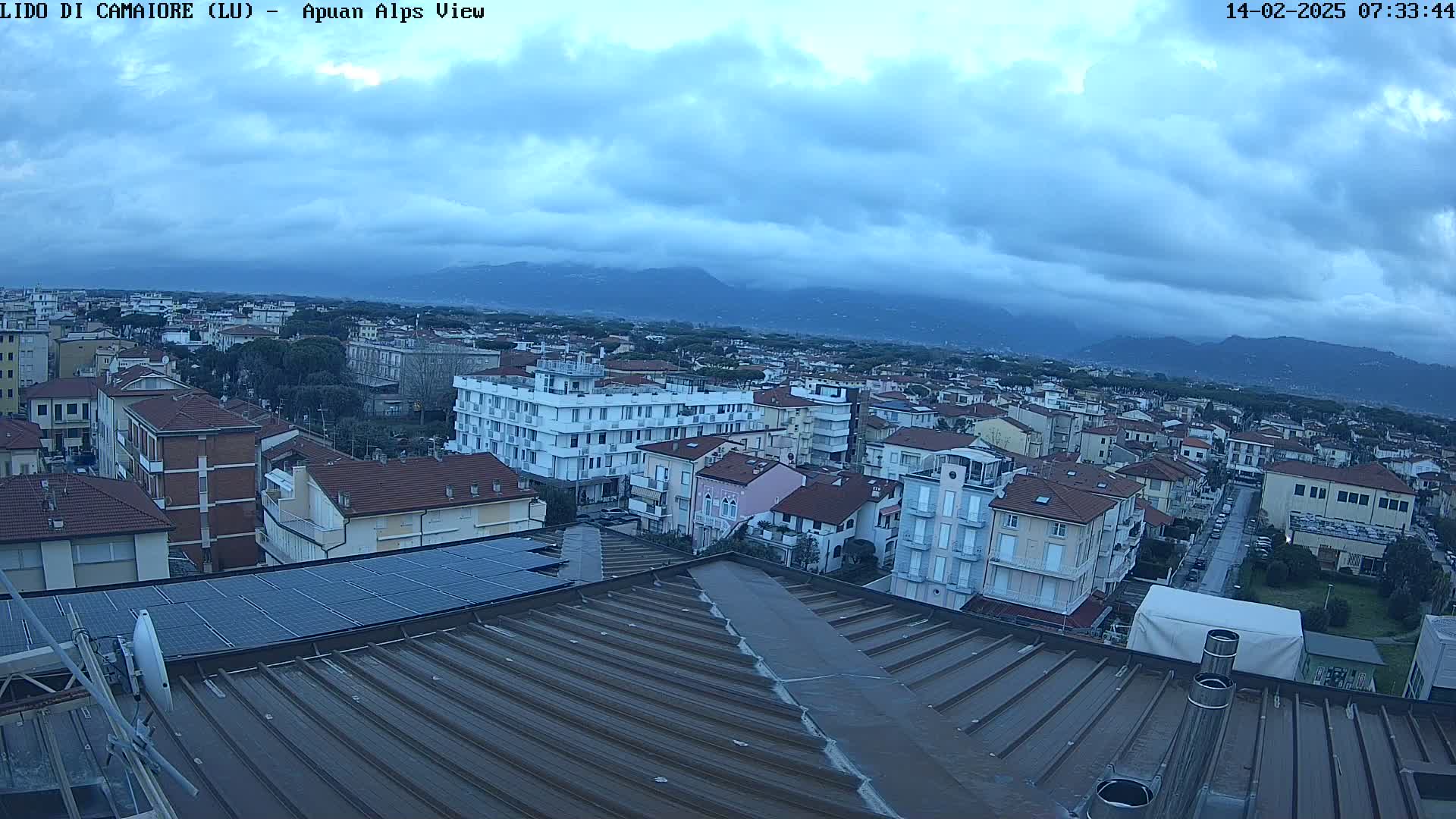 A view from a rooftop shows a town under a cloudy sky, with mountains visible in the distance.
