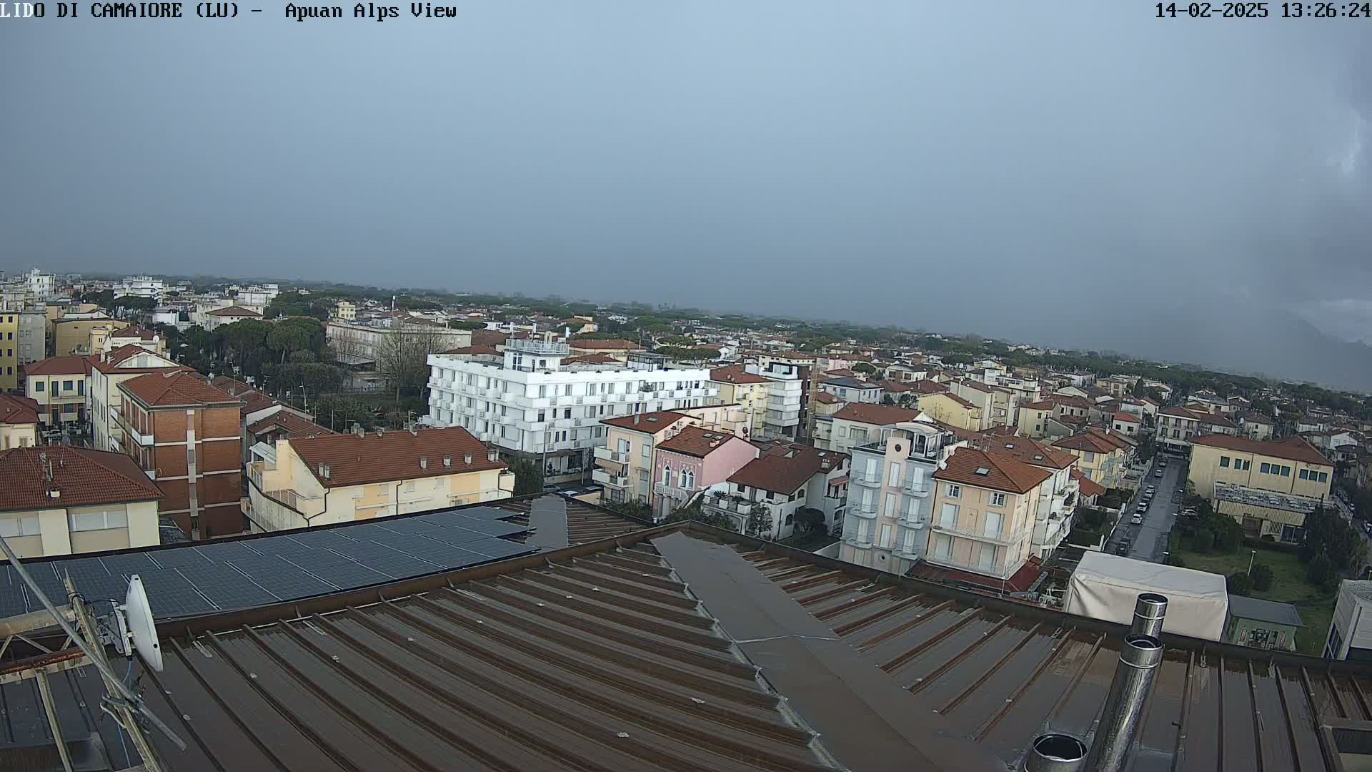 A wet, brown corrugated metal rooftop overlooks a town of low-rise buildings under an overcast sky.