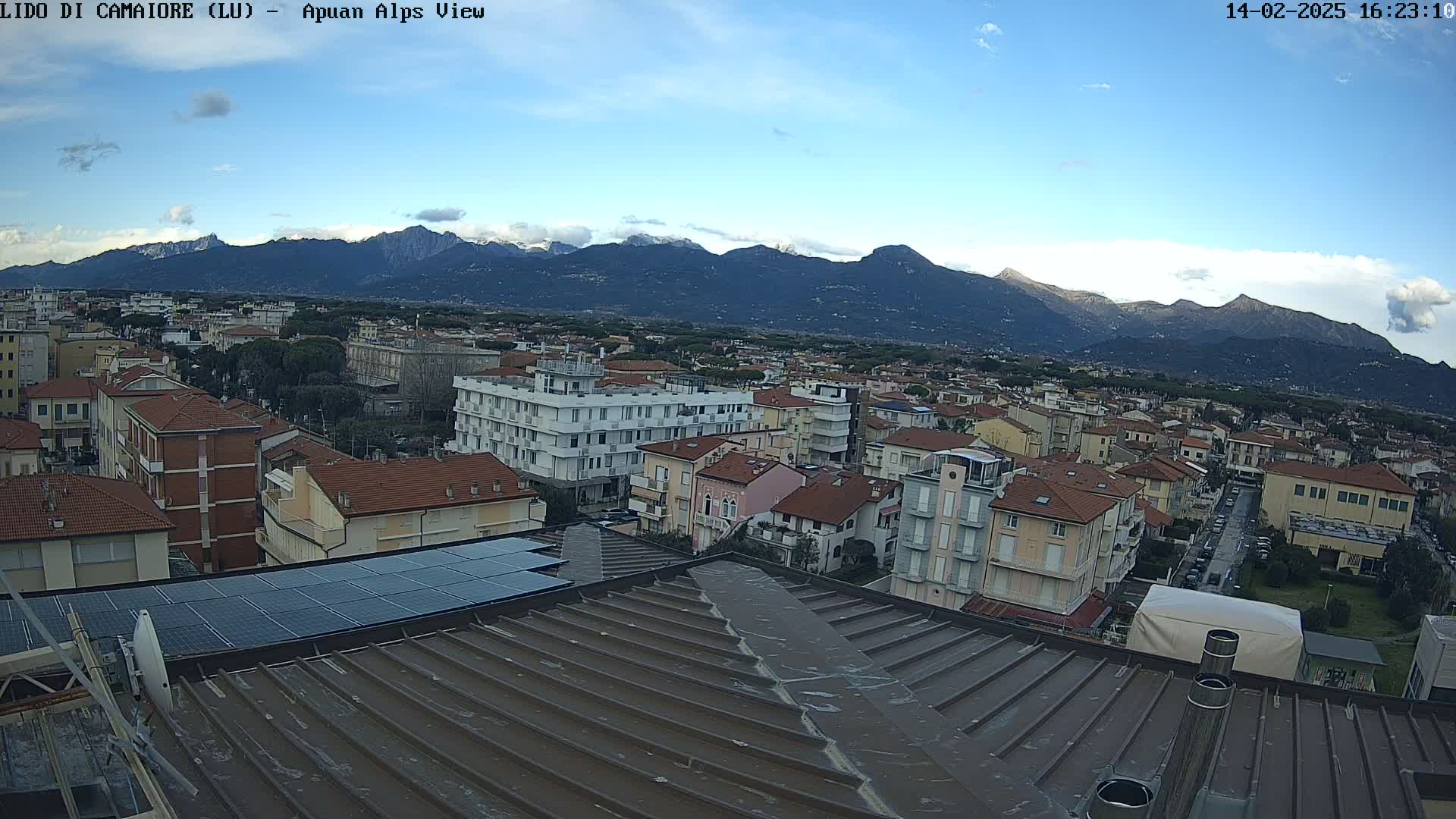 A rooftop view shows a town with multi-story buildings under a mostly clear blue sky, with snow-capped mountains in the distance.