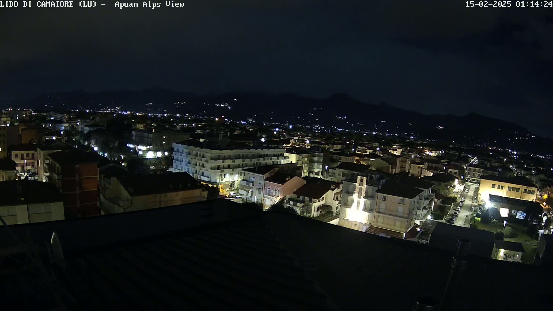 A nighttime aerial view of a town, illuminated by streetlights, with dark mountains in the background under a clear, dark sky.