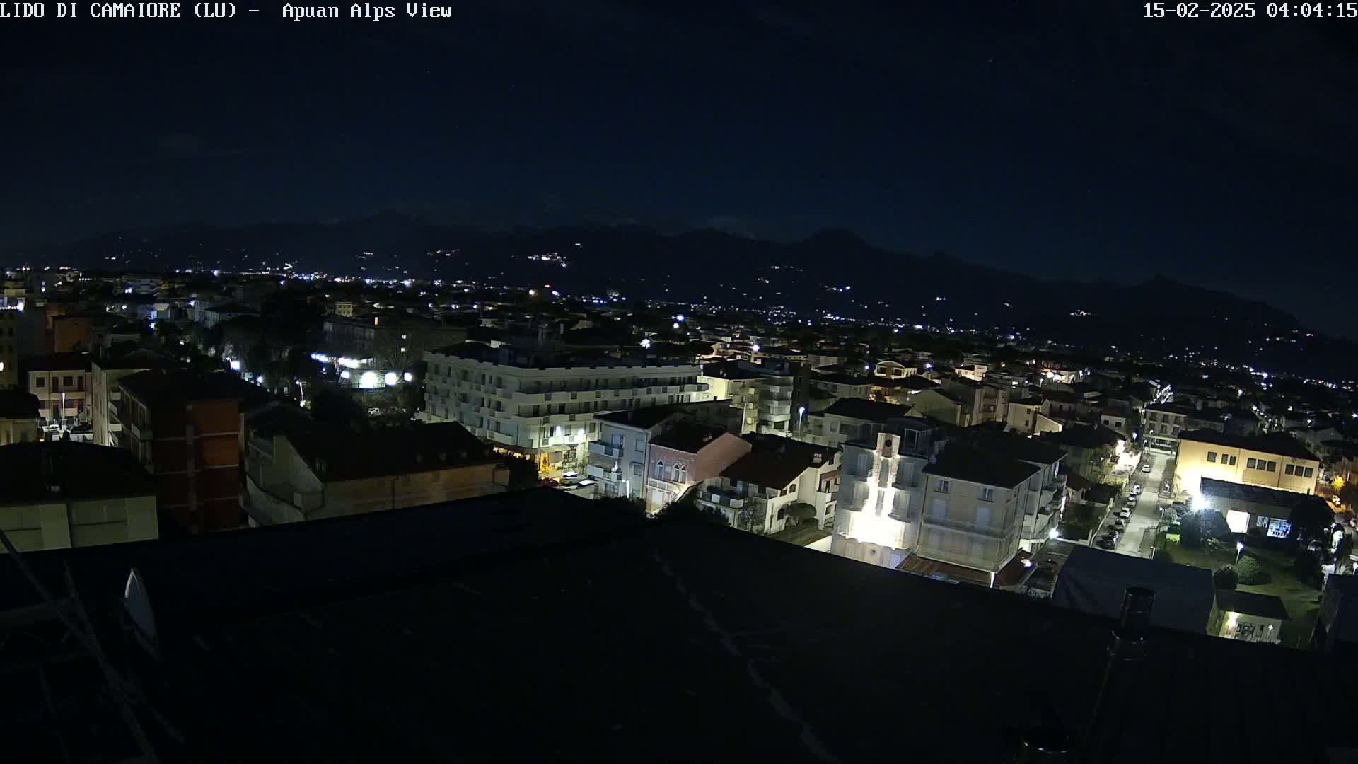 A nighttime aerial view of a town nestled below mountains, with dimly lit buildings and streets.