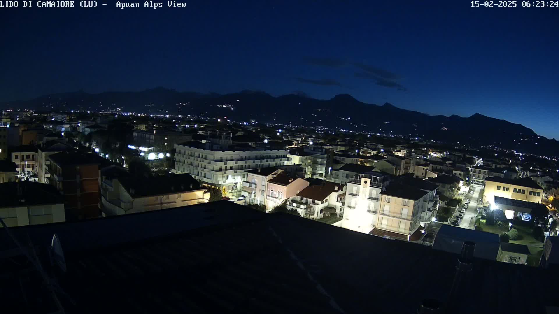 A nighttime aerial view shows a town illuminated, nestled against a dark mountain range under a clear, dark sky.