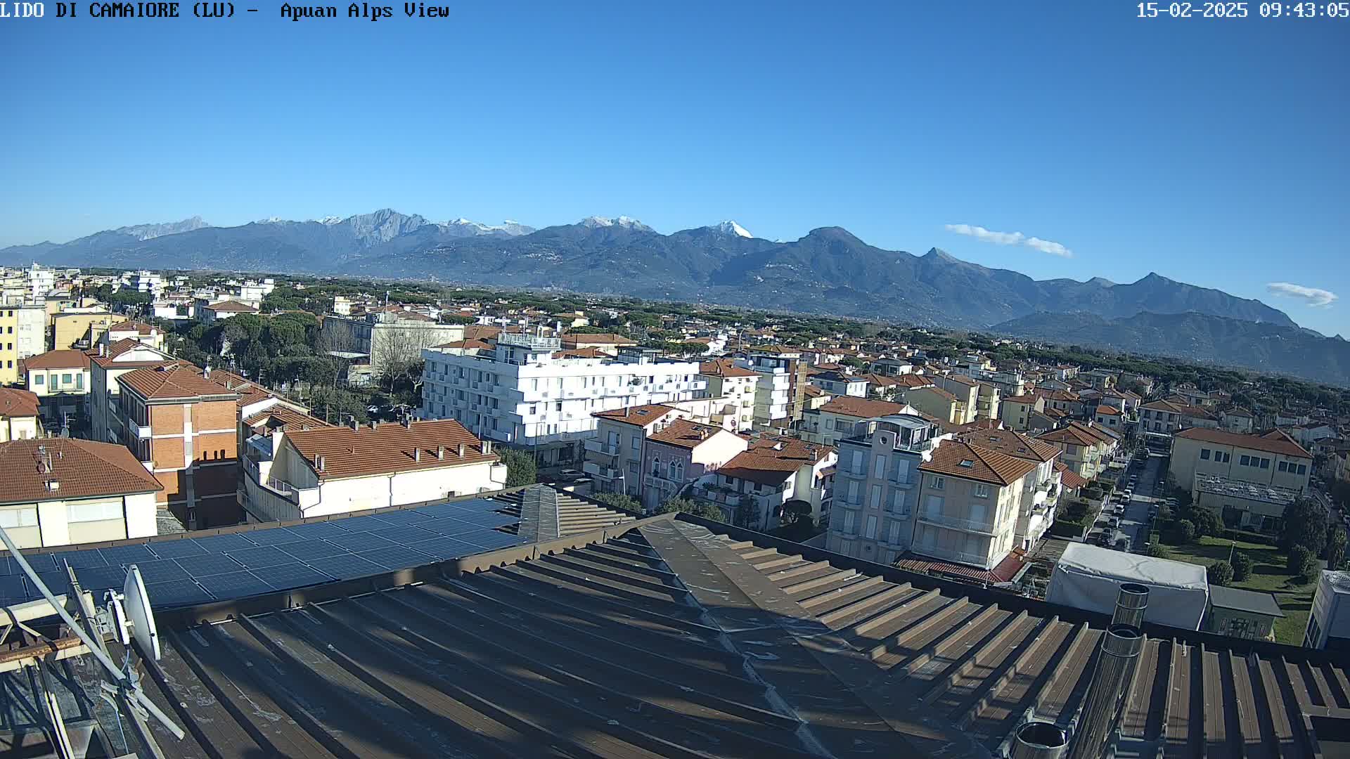 A clear, sunny day shows a rooftop view of a town nestled against a backdrop of snow-capped mountains.